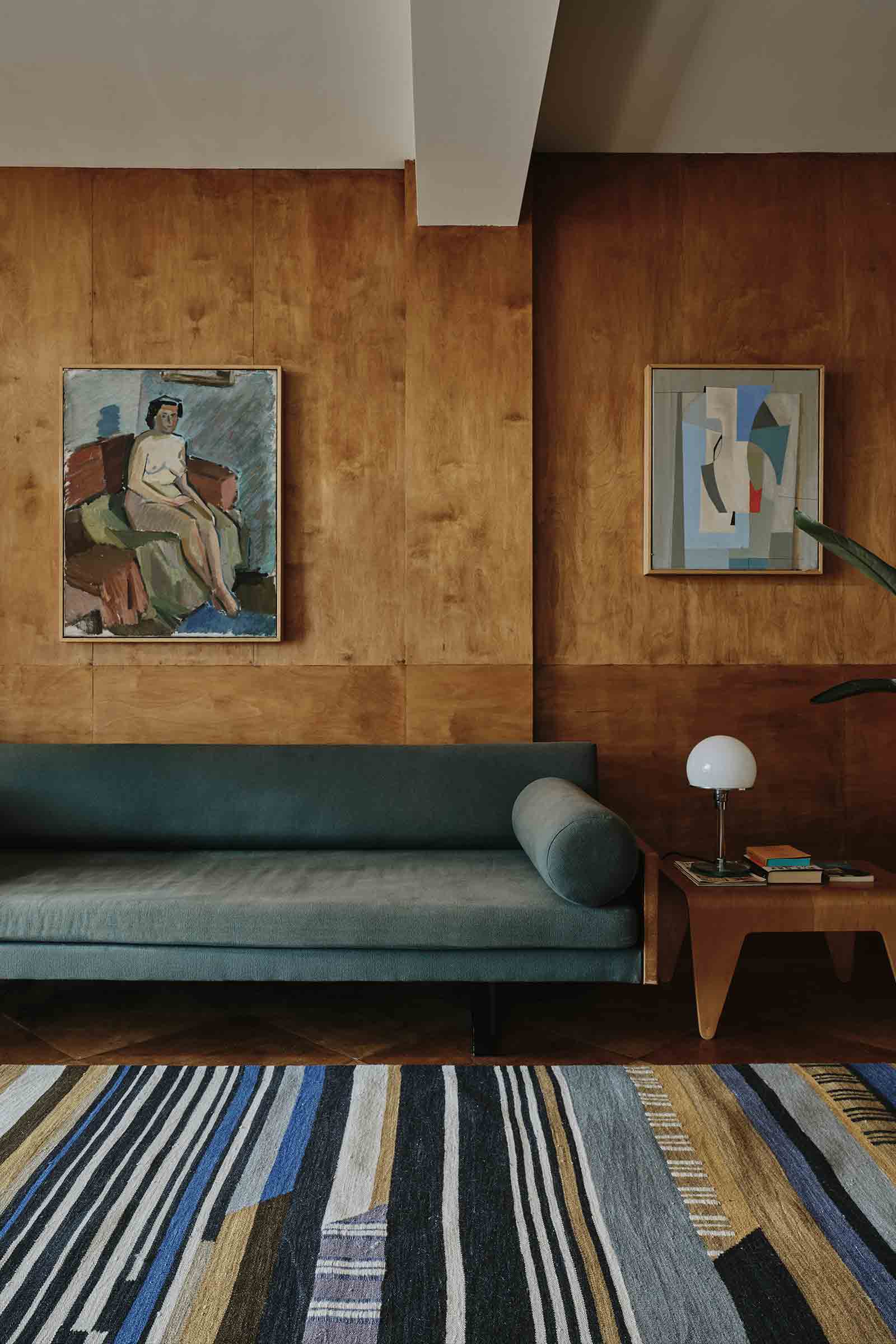 Dunkel gestreifter Bauhaus Teppich, der in einem Raum mit Holzwänden, an denen Bilder hängen, vor einem langen Sofa liegt