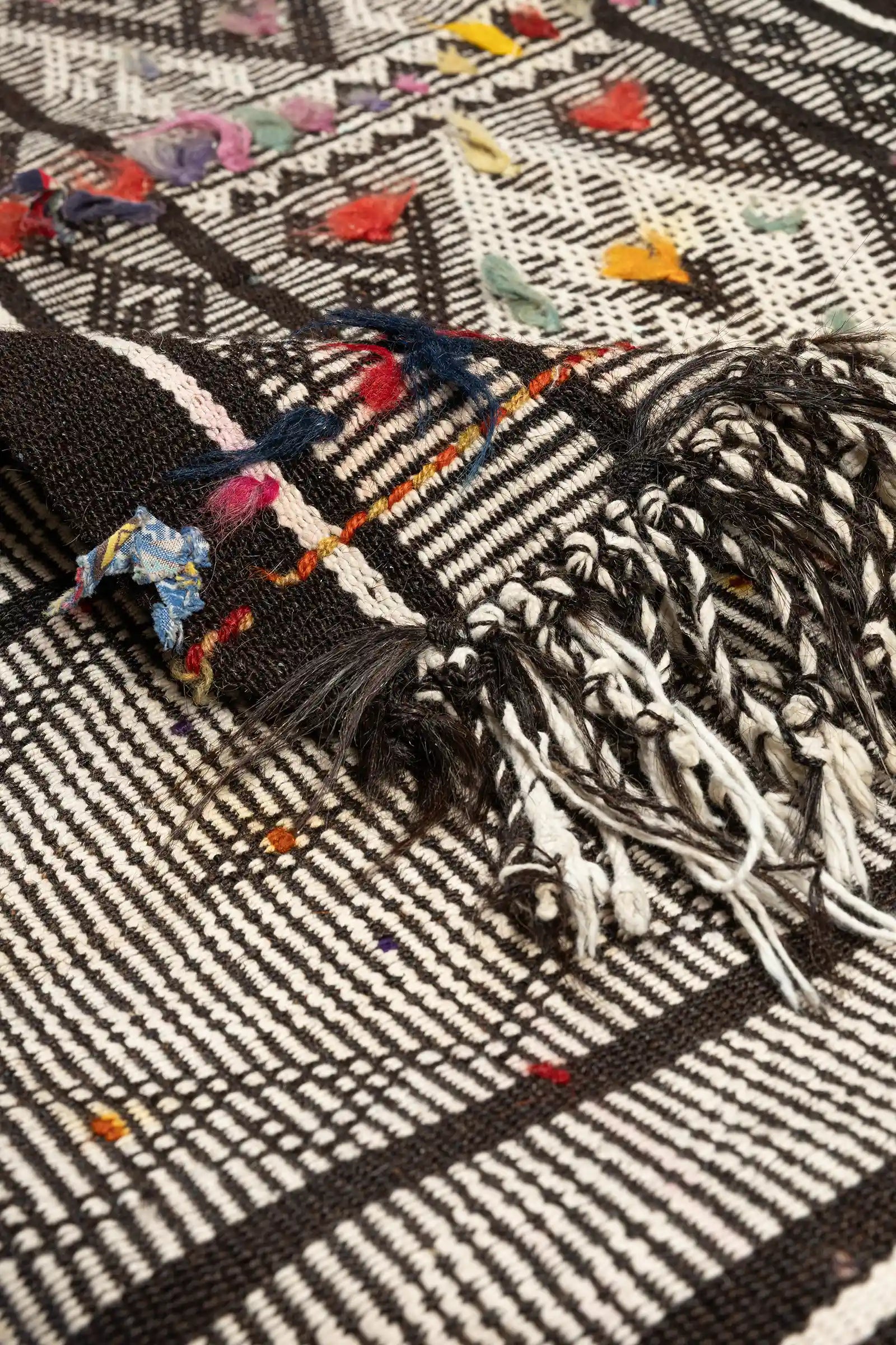 Teilansicht mit umgeschlagener Ecke des Kilims, die Vorderseite mit kräftigem Muster, die Rückseite zeigt die farbige Webkante und locker gebundene Fransen in Schwarz und Weiß.