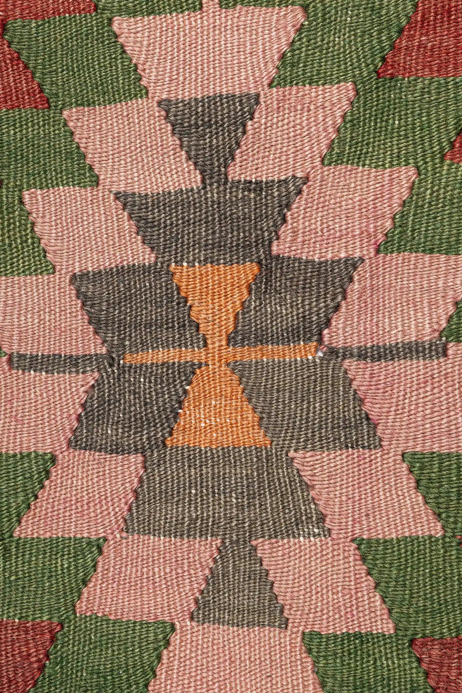 Nahaufnahme einer Raute mit rosafarbenem Zentrum, umgeben von abwechselnden Zacken in Grün, Hellblau, Rot und Violett, eingefasst von einer Violett-Orangen Bordüre.