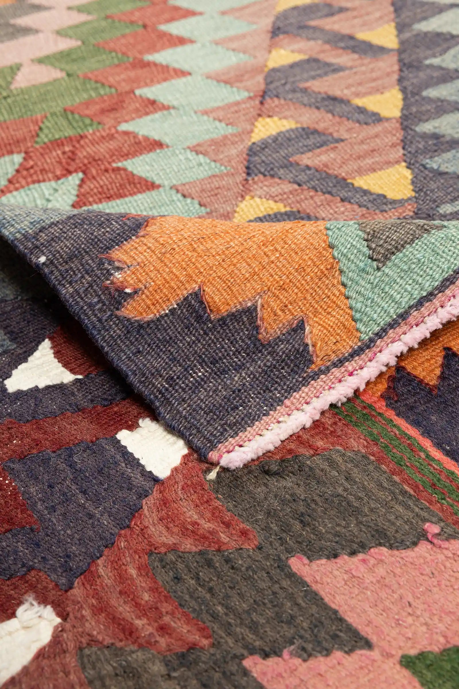 Detailansicht der umgeschlagenen Ecke des Kilims mit sichtbarer Rückseite in invertierten Farben und eingewebter Abschlusskante; kurze Fransen an der Teppichkante, farbliche Kontraste klar sichtbar.