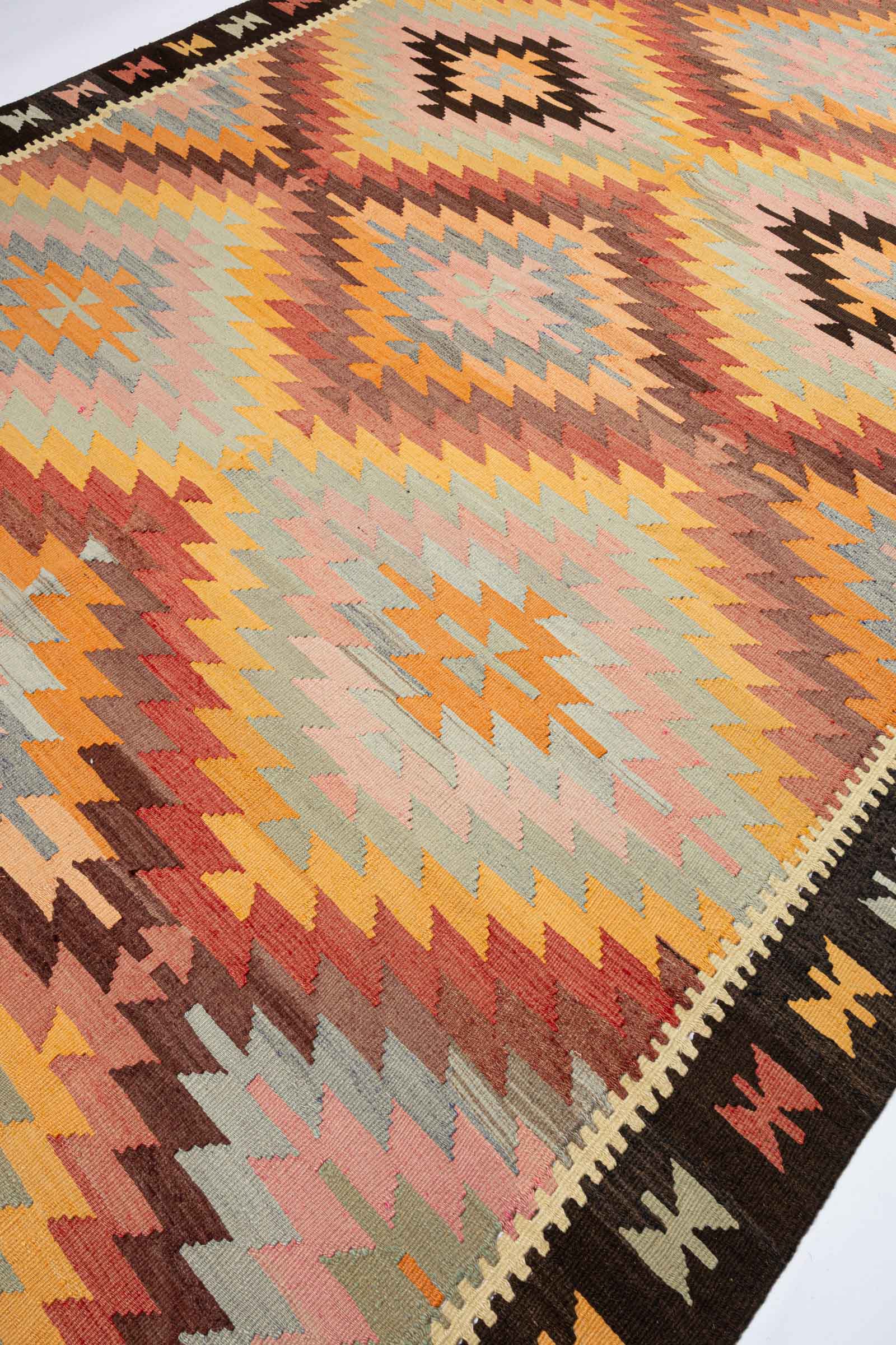 Diagonale Seitenansicht mit mehrfarbigen geometrischen Mustern in Orange, Rosa, Hellgrün und Braun, die sich über die Breite des Kelims erstrecken.