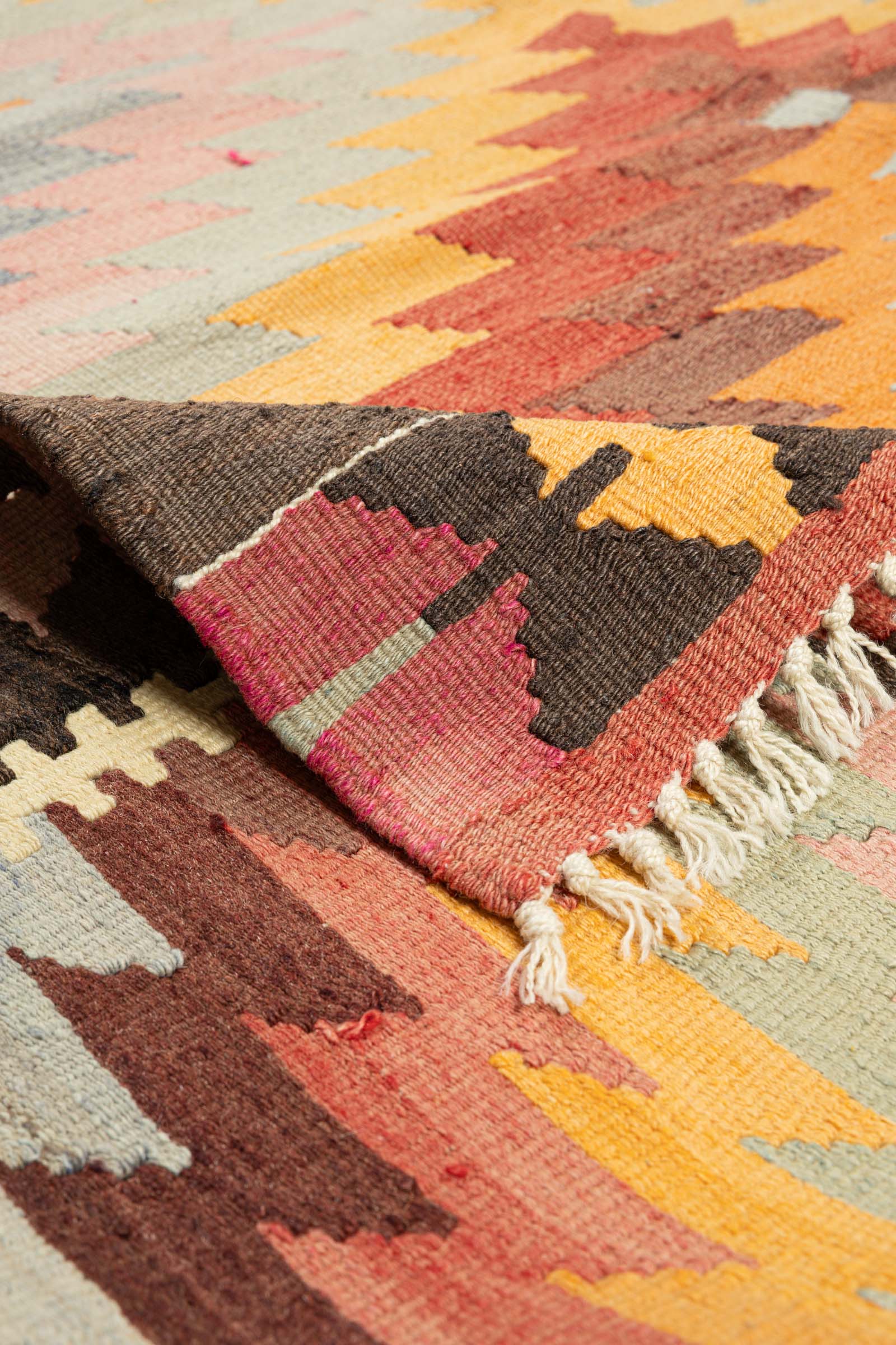 Umgeschlagene Ecke des Kelims mit sichtbarer Rückseite in denselben Farbtönen wie die Vorderseite; kurze, helle Fransen am Rand klar erkennbar.