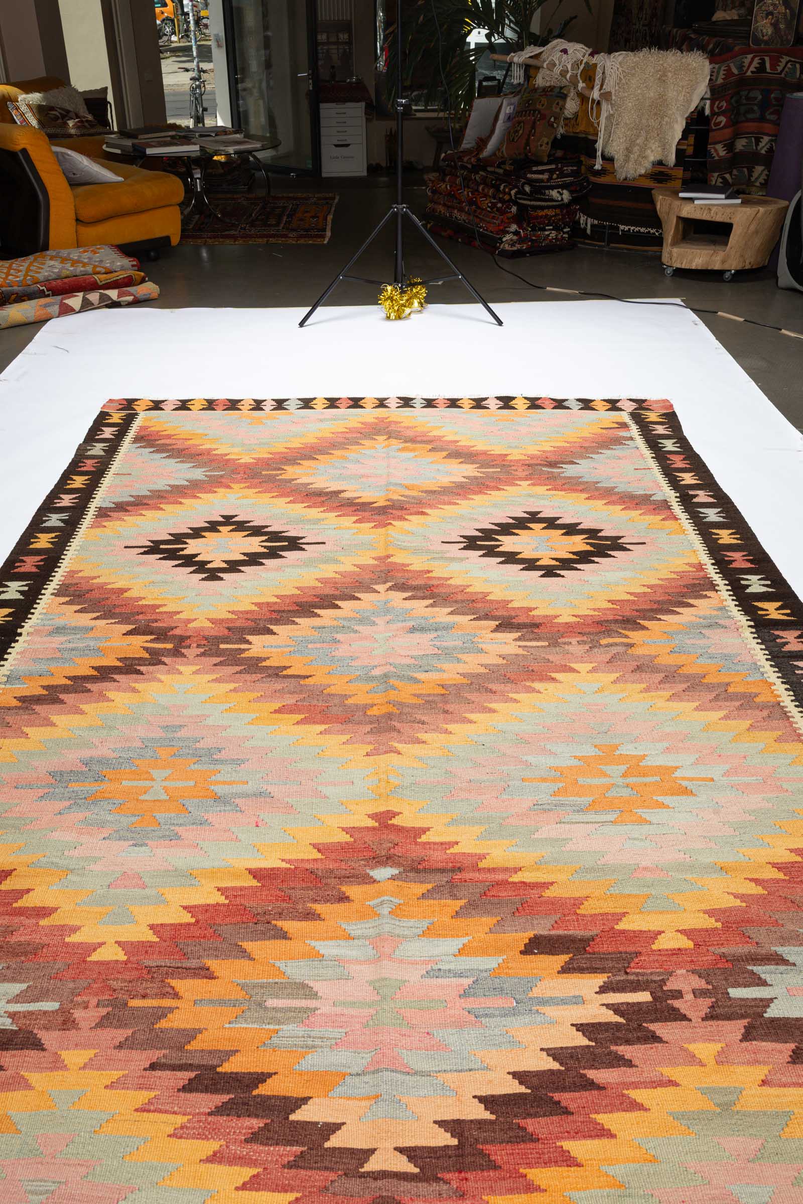 Schräg fotografierte Gesamtfläche des Kelims mit zentrisch verlaufenden Rauten aus zackigen Linien in Hellrosa, Türkis, Senfgelb und Braun, die Tiefe erzeugen.