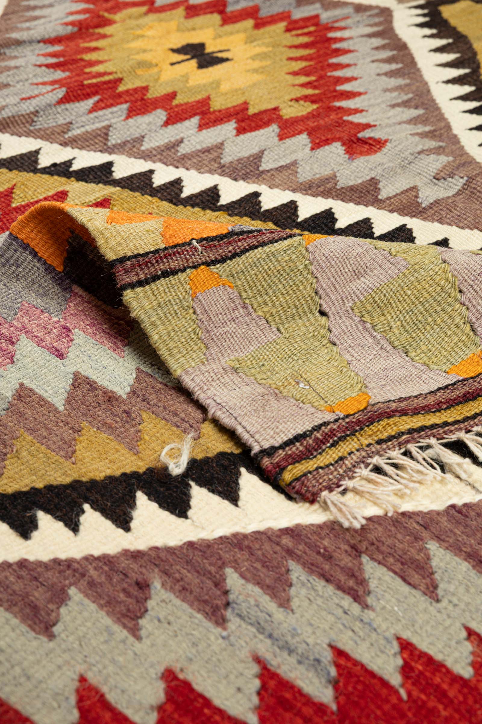 Umgeschlagene Ecke des Kelims mit sichtbarer Rückseite in ähnlicher Farbgebung; kurze, cremefarbene Fransen am unteren Rand sichtbar.