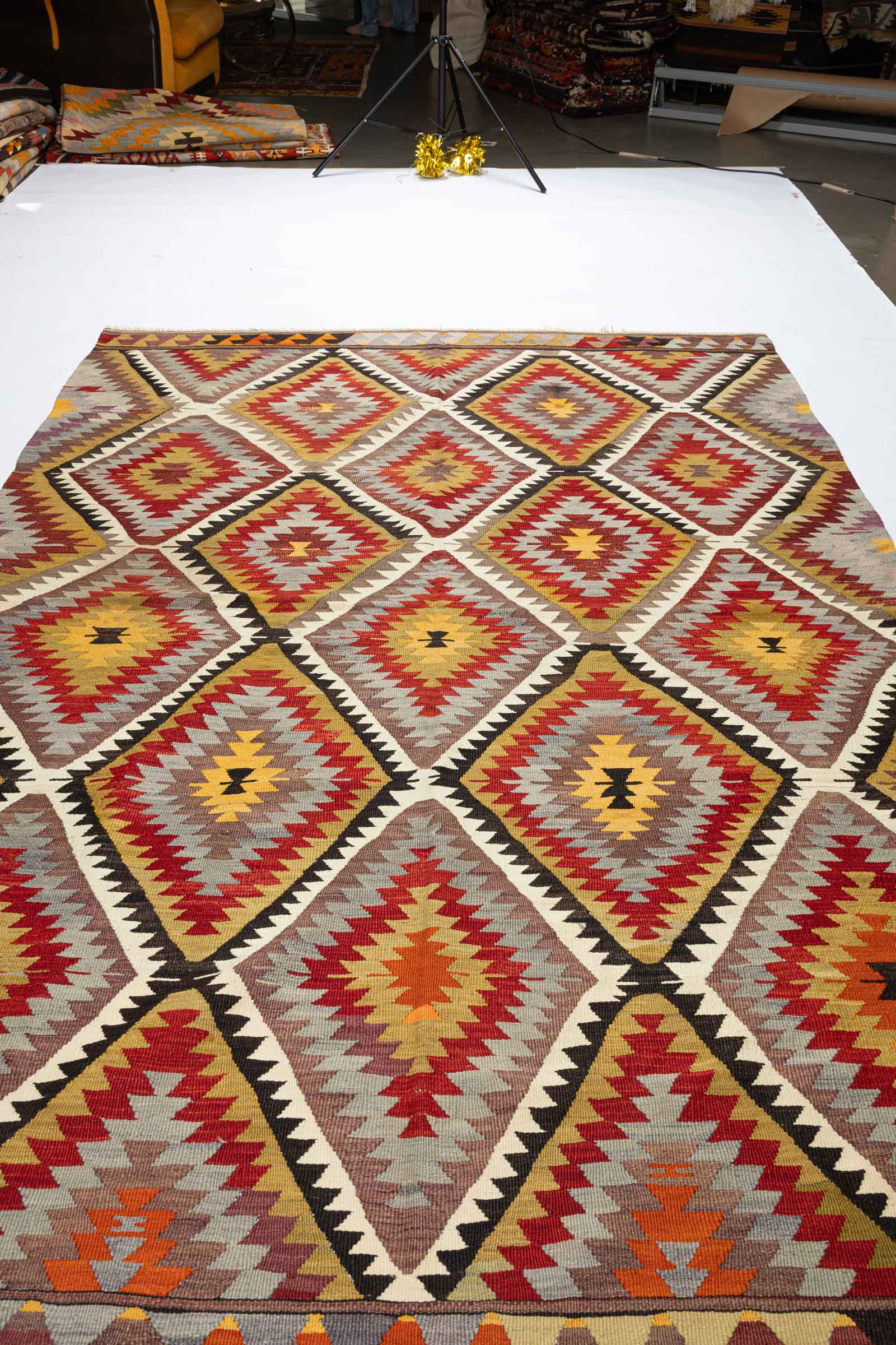 Schräg aufgenommene Gesamtfläche des Kelims mit dichter Reihung von symmetrischen Zackenrauten, deren Farben sich in Rot, Braun, Grau und Gelb abwechseln.