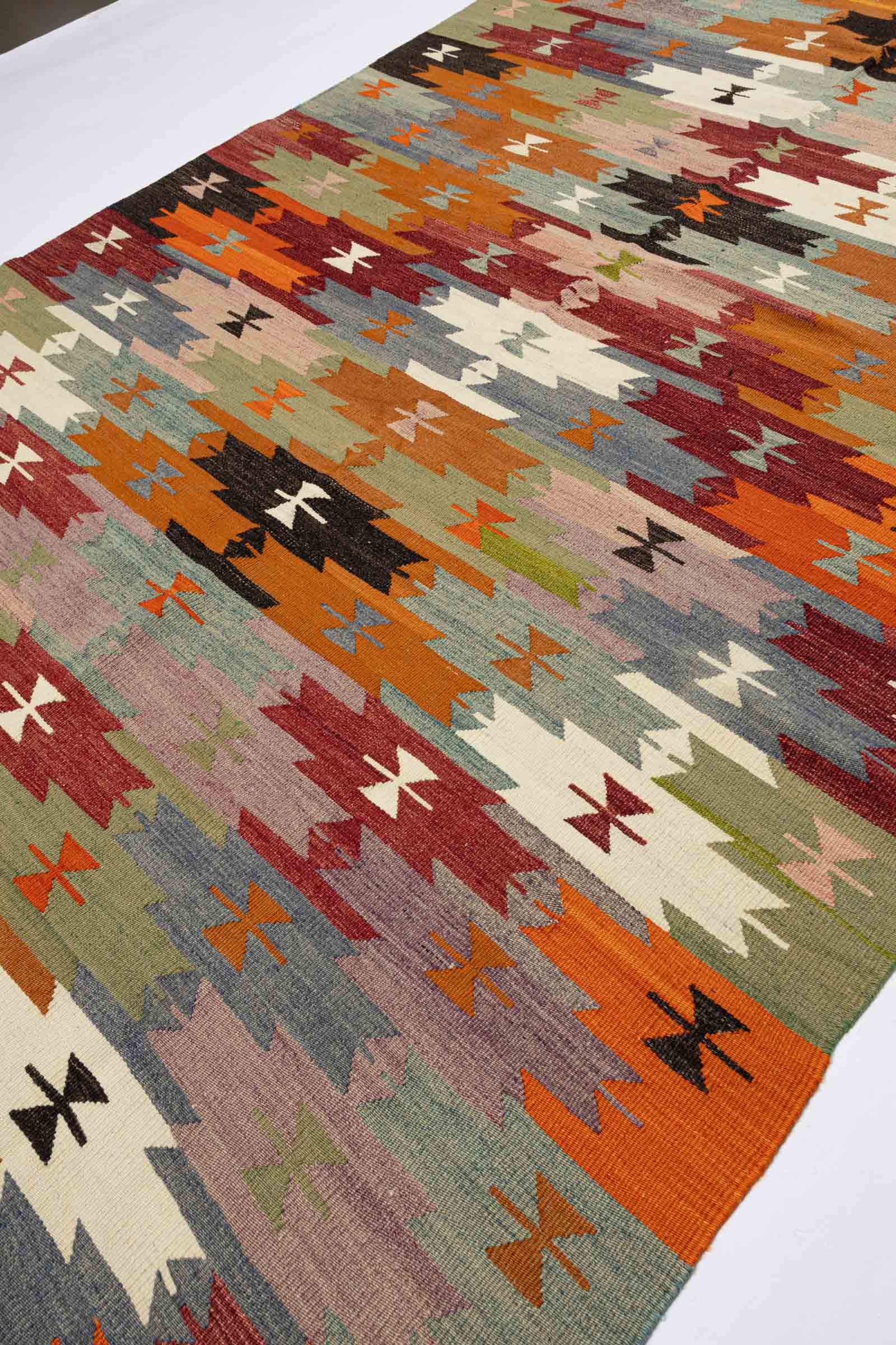 Diagonale Seitenansicht des Kilims mit versetzten Streifen und geometrischen Symbolen in wechselnden Farbkombinationen wie Rostrot-Weiß, Schwarz-Orange und Grün-Rosa auf einem hellen Fotohintergrund.