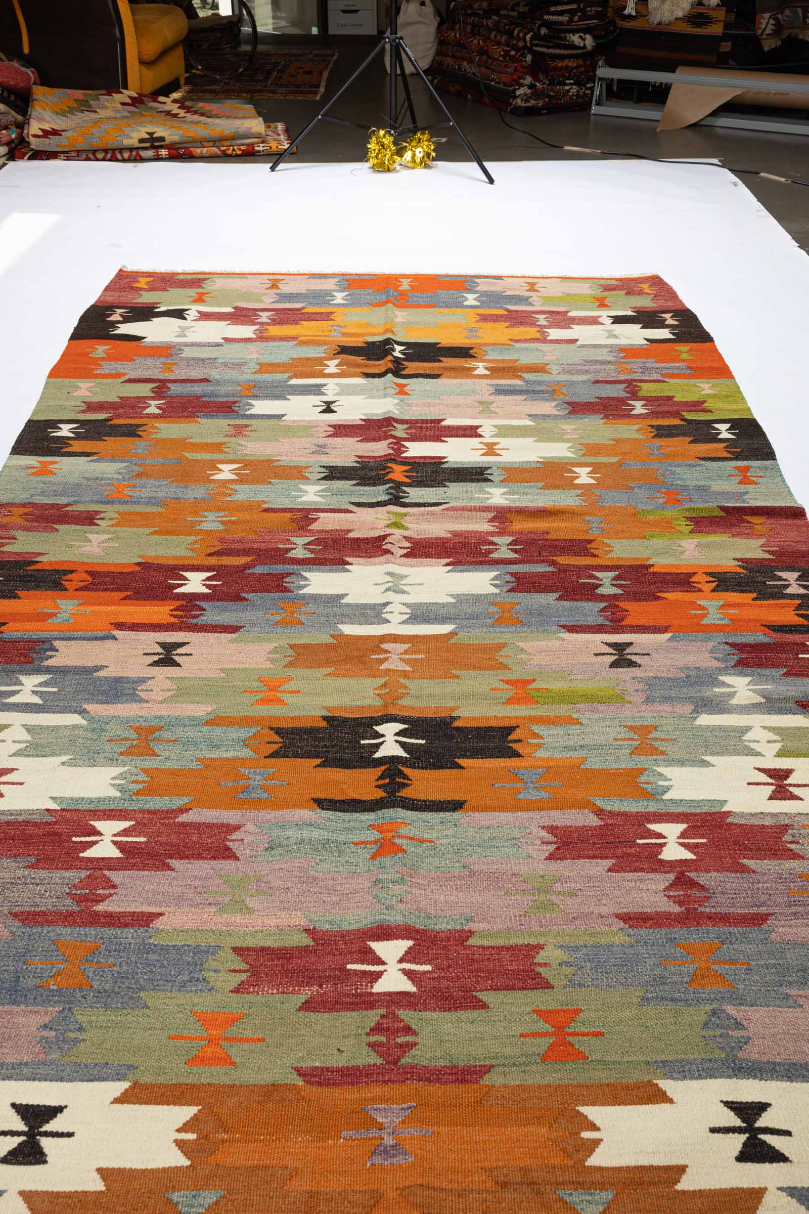 Der flach gewebte Vintage-Kelim aus schrägem Winkel aufgenommen, mit horizontalen Reihen in leuchtenden Farben wie Orange, Violett, Schwarz und Grün, durchsetzt mit farblich kontrastierenden geometrischen Mustern.