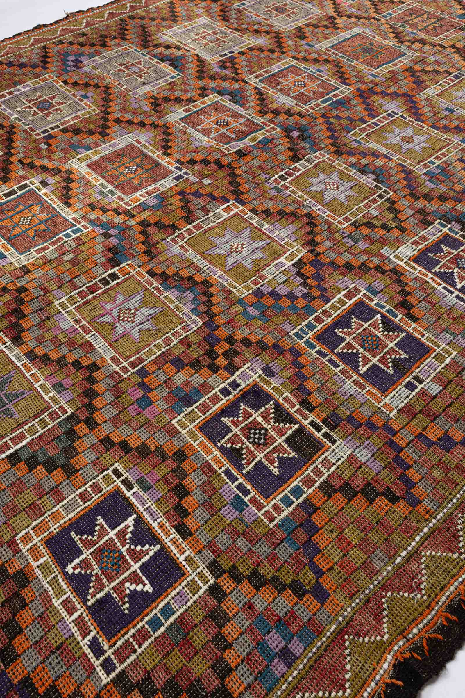 Seitliche Nahaufnahme der eingewebten Karomuster in Schwarz, Orange, Lila, Grün und Rot, unterbrochen von weißen Linien, die die sternförmigen Rechteckfelder geometrisch einfassen. Die Textur des dickeren Garns ist deutlich erkennbar.