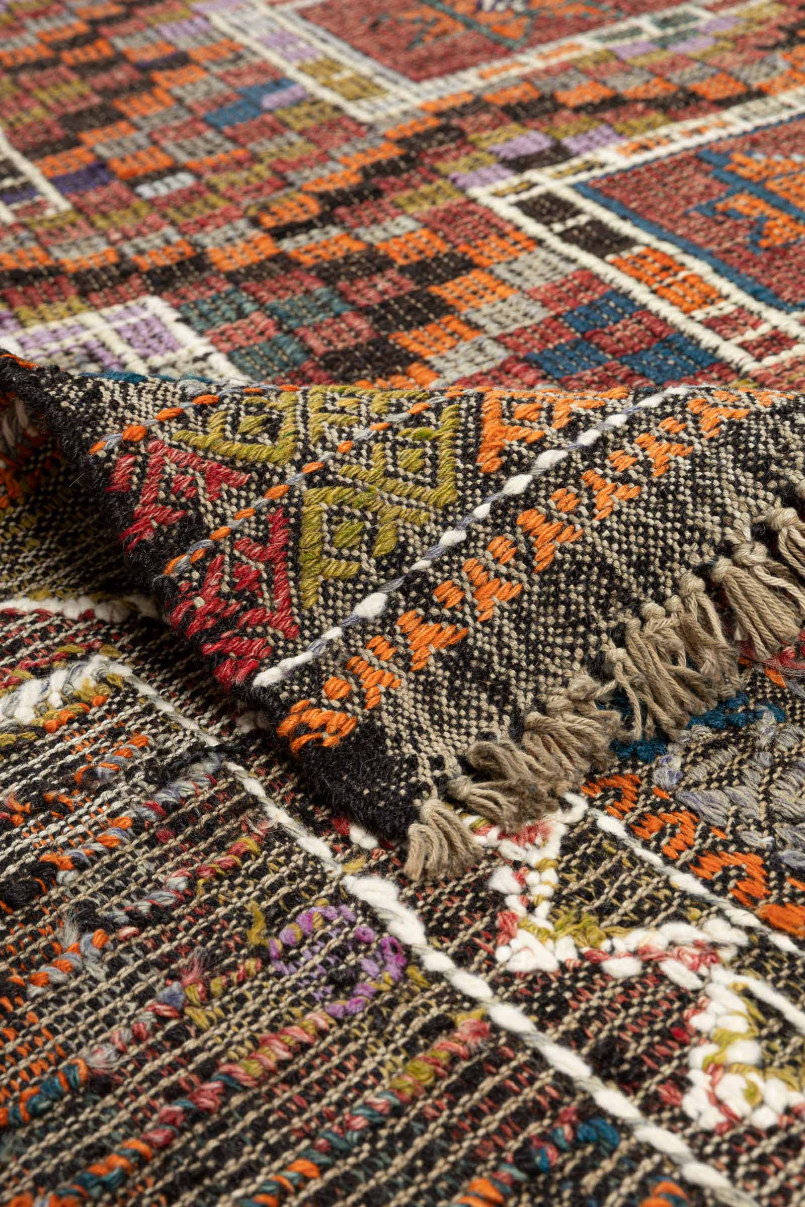 Ecke des Kelims leicht umgeschlagen, um die Rückseite mit dunklem Grundton und mehrfarbig eingewebten geometrischen Mustern in Rot, Oliv, Orange und Gelb zu zeigen. Die helle Fransenreihe verläuft entlang der unteren Kante.