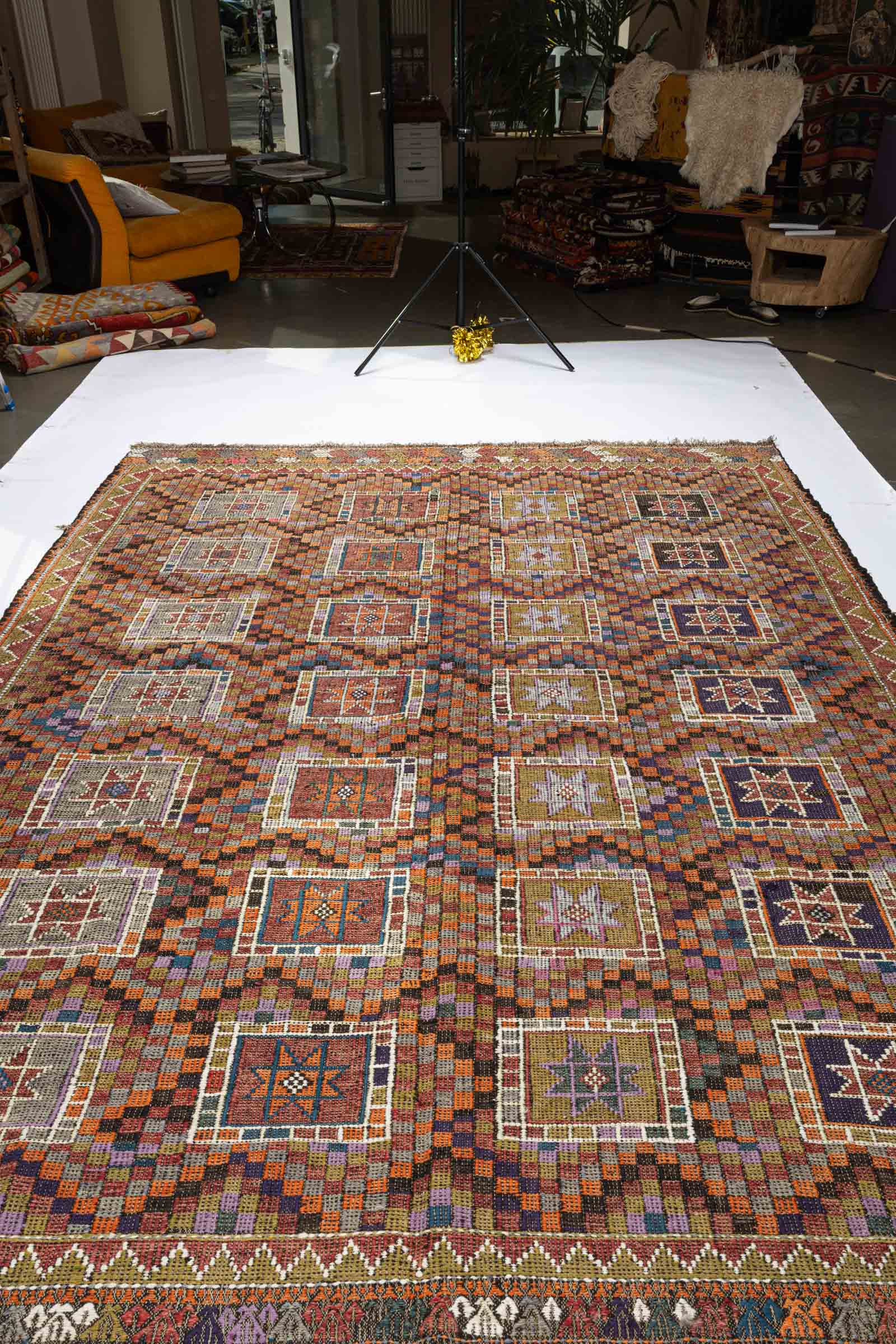 Schräg aufgenommene Aufnahme des Kelims mit sich wiederholenden sternförmigen Motiven in rechteckigen Feldern, die in dichten, bunten Karomustern eingebettet sind. Die Oberfläche zeigt sichtbare Wellen und leichte Strukturunregelmäßigkeiten.