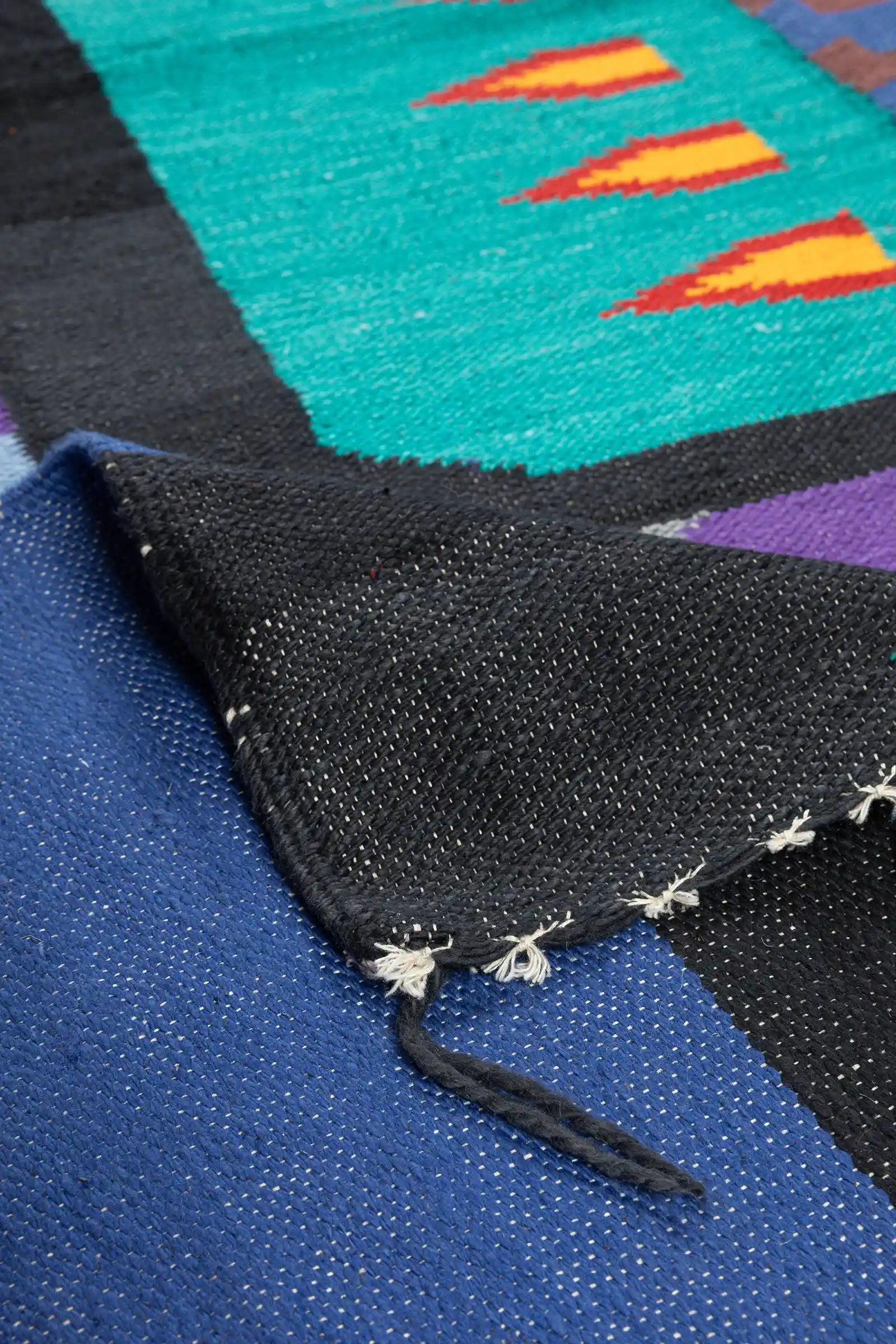 Ansicht einer umgeschlagenen Ecke des Kelims mit schwarzer Rückseite, weißem Fransenabschluss und sichtbarer Verarbeitung der farbigen Vorderseite.