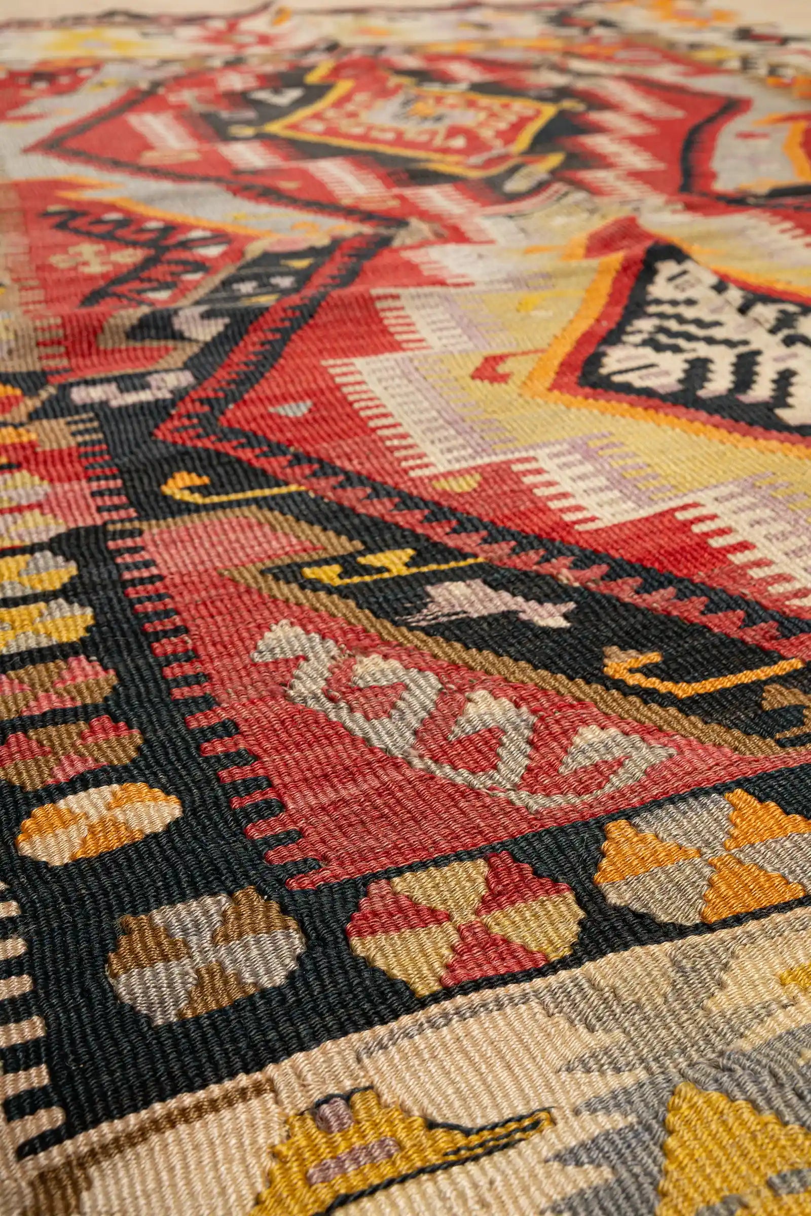 Makroaufnahme eines roten Rautelements mit eingewebten schwarzen und beigen Linien sowie einem gelbgrünen Kreuzmotiv in der Mitte. Die dichte Flachgewebe-Struktur ist fein und gleichmäßig.