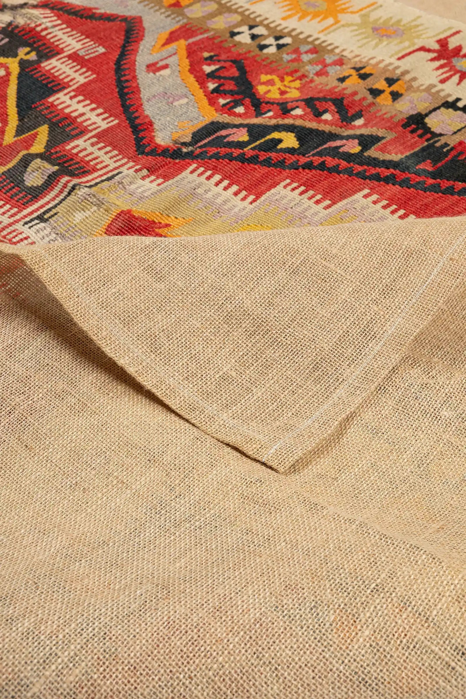 Ansicht einer leicht zurückgeschlagenen Ecke des Kelims, die die naturfarbene, grob gewebte Rückseite sowie die glatte Webvorderseite zeigt. Fransen entlang der Kante sind unregelmäßig und teilweise abgenutzt.