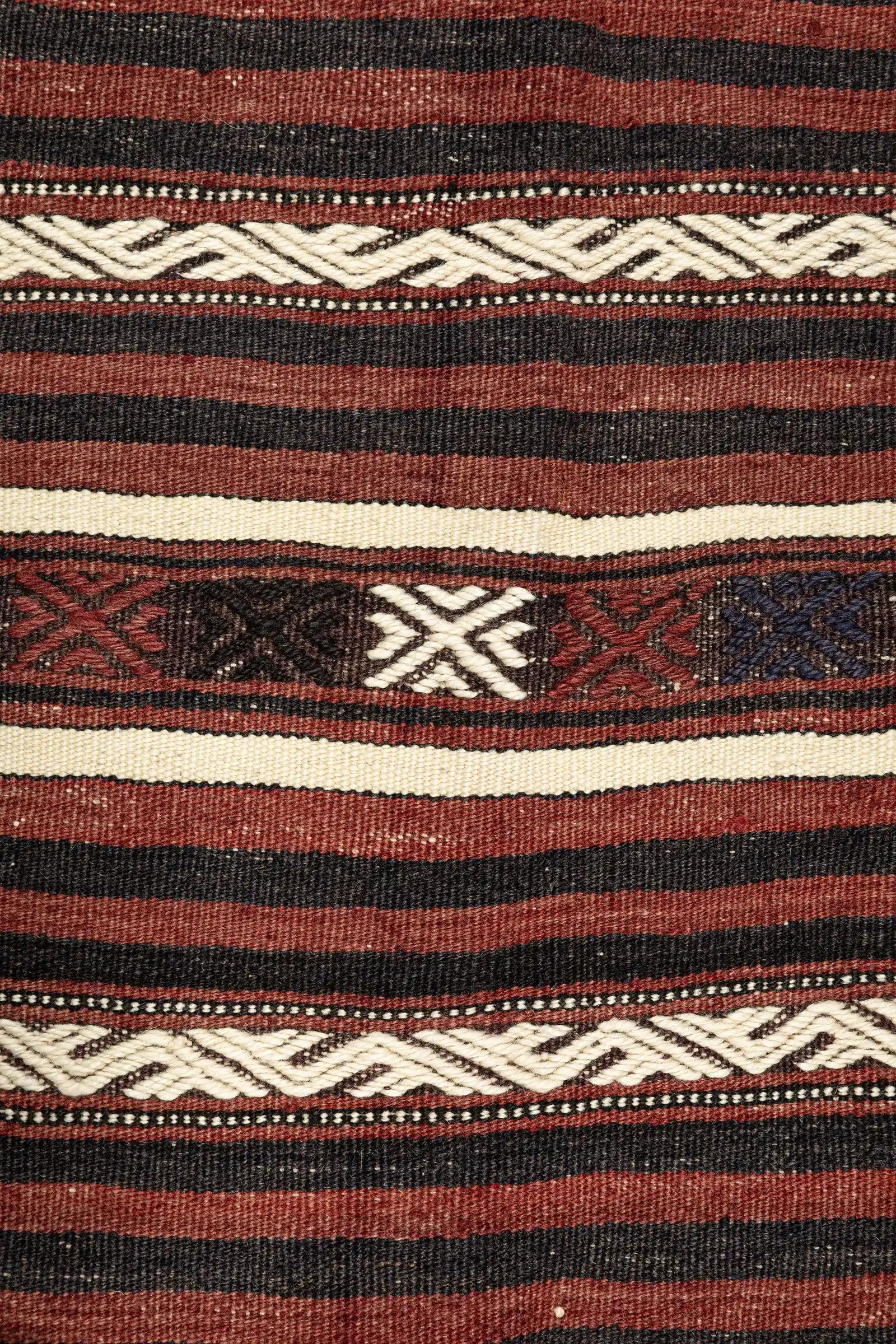 Detailaufnahme eines weißen Streifenelements – mittig ein cremefarbenes Rautenmuster auf dunklem Grund, flankiert von weiteren Rauten in Schwarz, Rot und Blau, eingefasst von schmalen Linien in Rotbraun und Schwarz