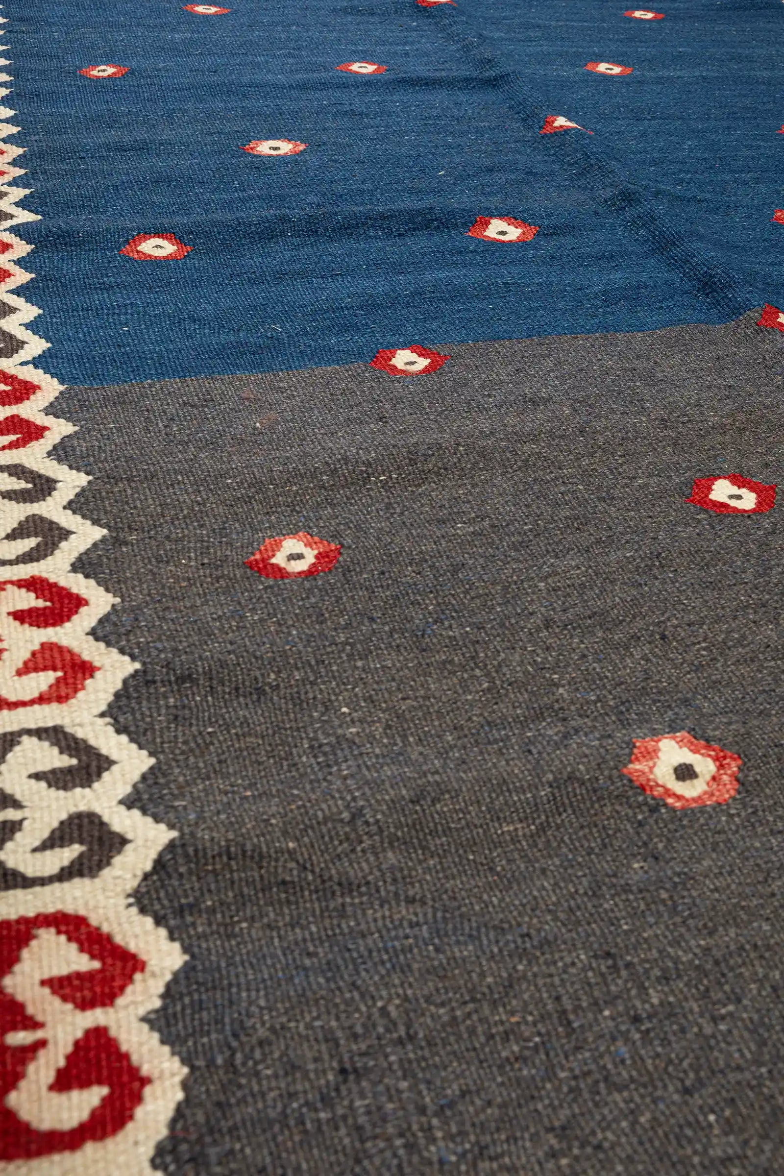 Texturdetail mit dichter Wollstruktur in indigoblauem Grund, einer kleinen roten Blüte mit hellem Zentrum und Teil der umlaufenden Bordüre in geometrischer Gestaltung.