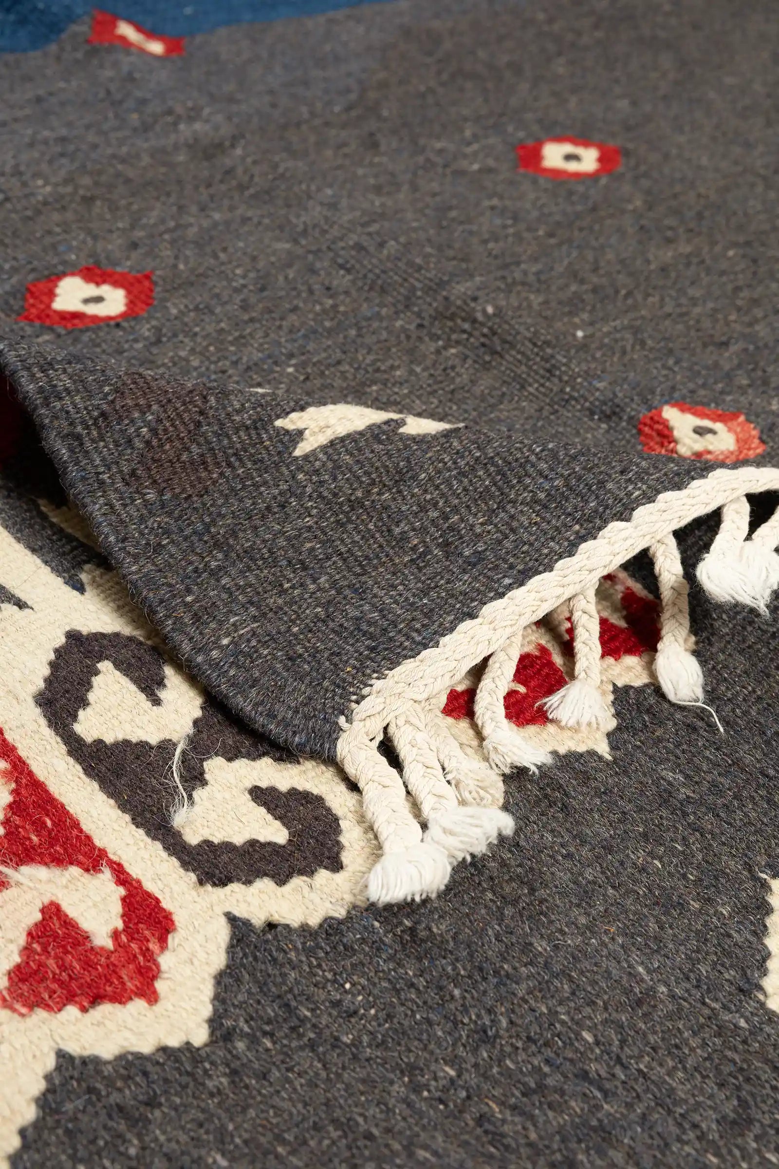 Umgeschlagene Ecke des Kelims zeigt die graue Rückseite, weiße Wollfransen sowie die kontrastreiche Bordüre mit roten und braunen Details.