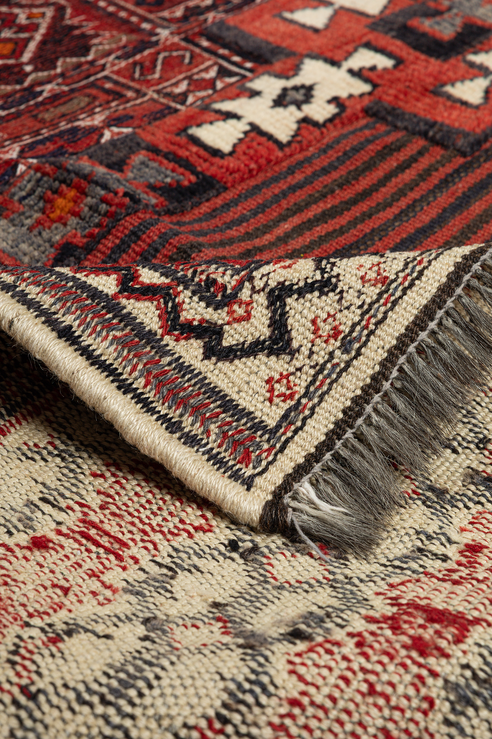 Ansicht der oberen rechten Ecke des Kilims mit nach hinten umgeschlagener Ecke. Sichtbar sind die Vorderseite mit rotem Streifenmuster und cremefarbener Bordüre sowie die Rückseite mit flach gewebtem Muster. Dunkle Fransen und saubere Kantenstruktur deutlich erkennbar.