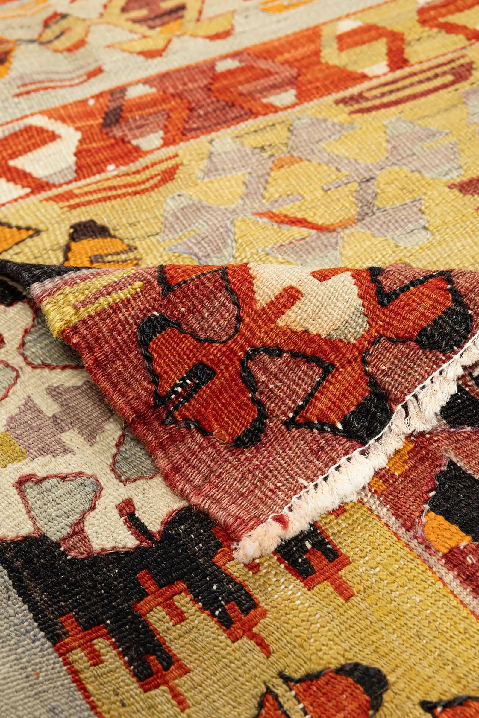 Bild zeigt eine Ecke des Kilims nach oben geklappt: Oben die Vorderseite mit geometrischen, ineinandergreifenden Mustern in Rot, Schwarz und Beige; unten die Rückseite mit gut erkennbarer Webstruktur, Fransenansatz und Farbdurchdringungen in Ziegelrot und Orange.