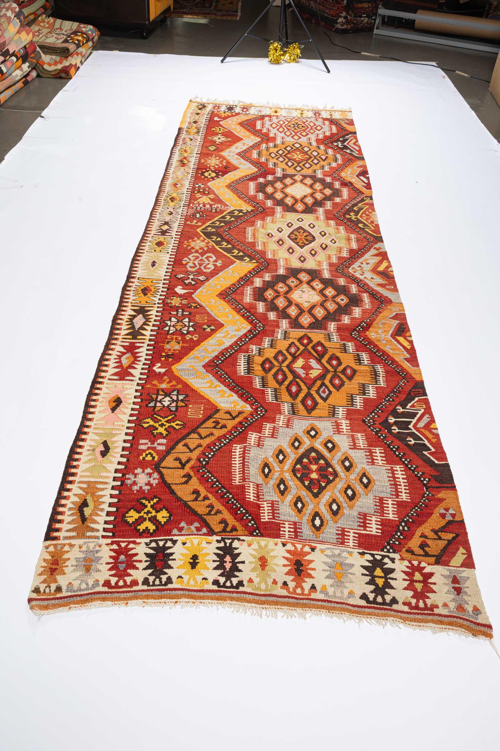 Diagonal aufgenommene Teilansicht des Kelims mit mehreren ineinander übergehenden Rautenmustern in Rot, Orange, Beige und Dunkelbraun sowie gelb-weißen Randornamenten.