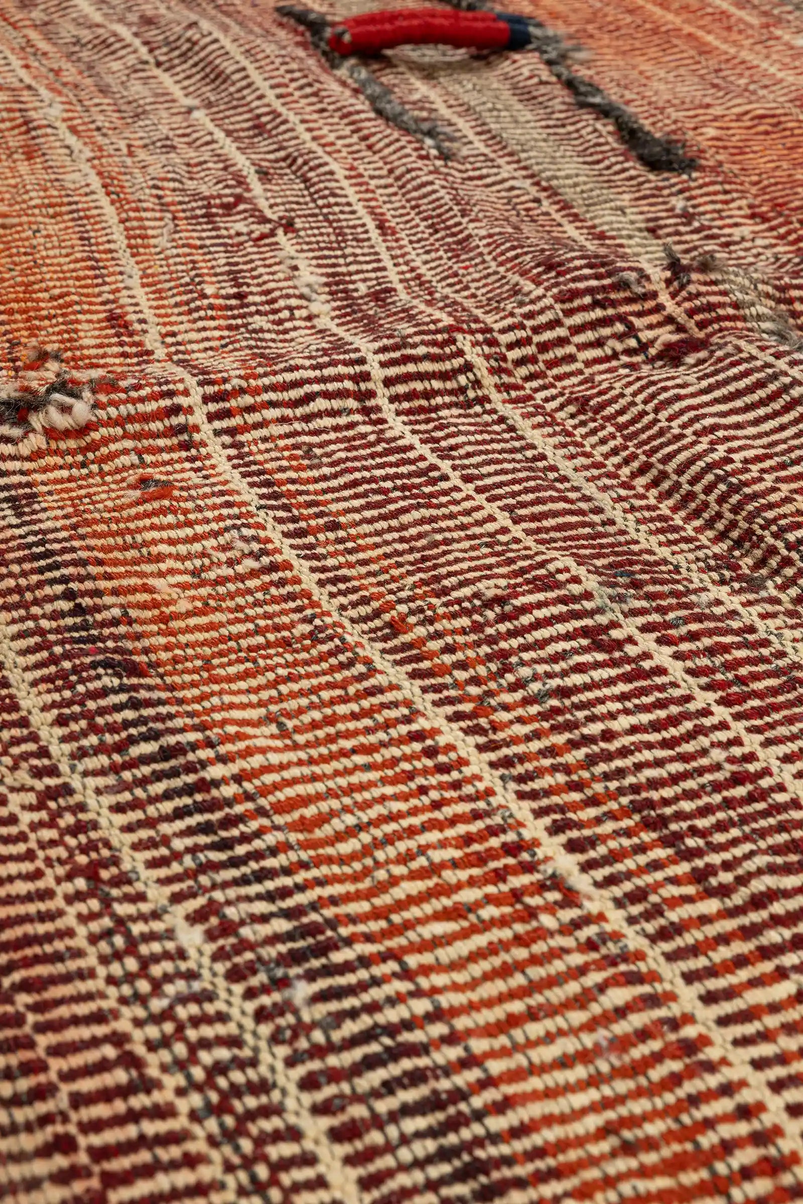 Detailausschnitt der dichten, flach gewebten Oberfläche mit rot-beigen Streifen, ergänzt durch dunkelgraue Garnapplikationen. Im Zentrum ein dekoratives Seil mit dichter Umwicklung aus rotem und blauem Wollgarn.