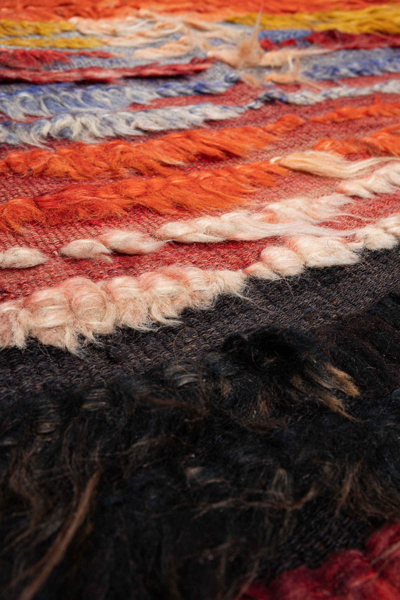 Sehr naher Blick auf die Oberfläche des Kilims mit detaillierter Sicht auf gelbe, orangefarbene und hellrosa Wollfasern, die aus dem grob gewebten blaugrauen Hintergrund herausragen.