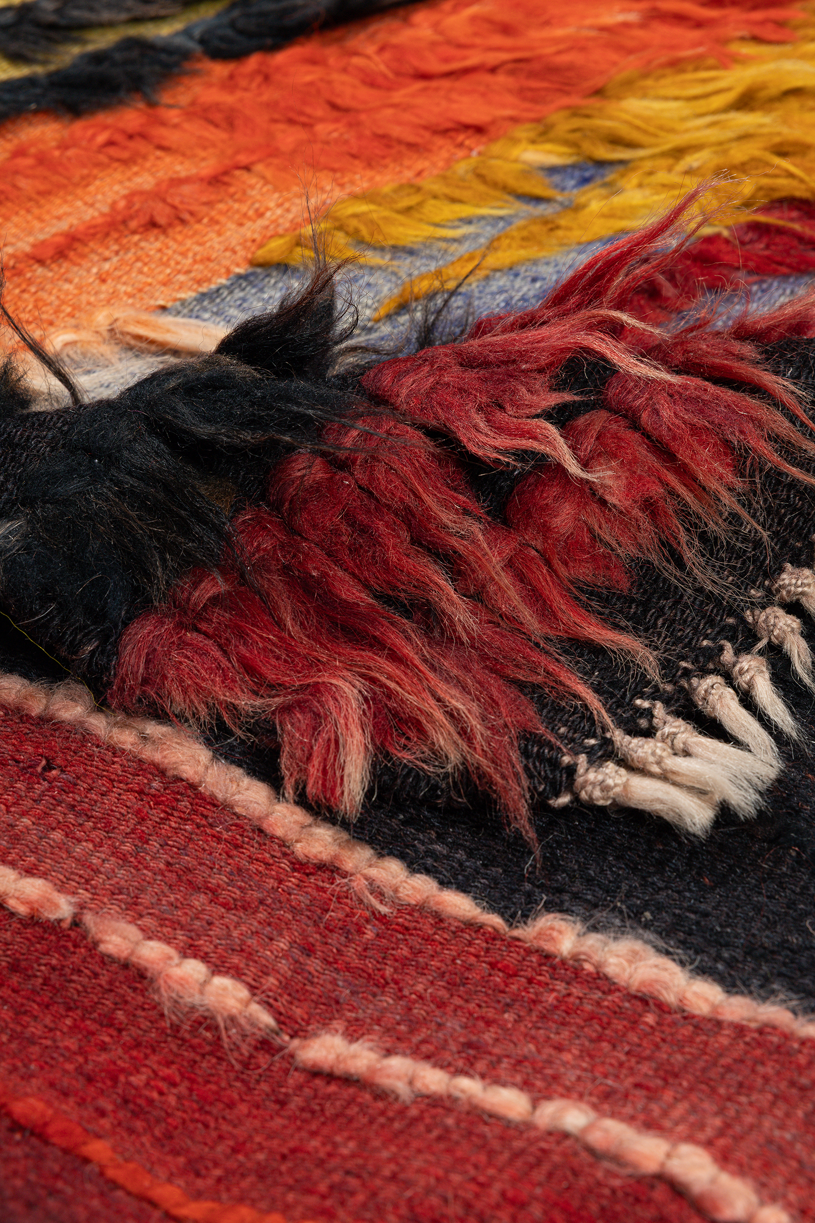 Aufnahme der Ecke des Kilims mit nach oben umgeschlagener Kante; sichtbar sind die kurzen, weißen Fransen sowie die Unterseite mit gewebter Struktur in Beige, Schwarz und Rot.