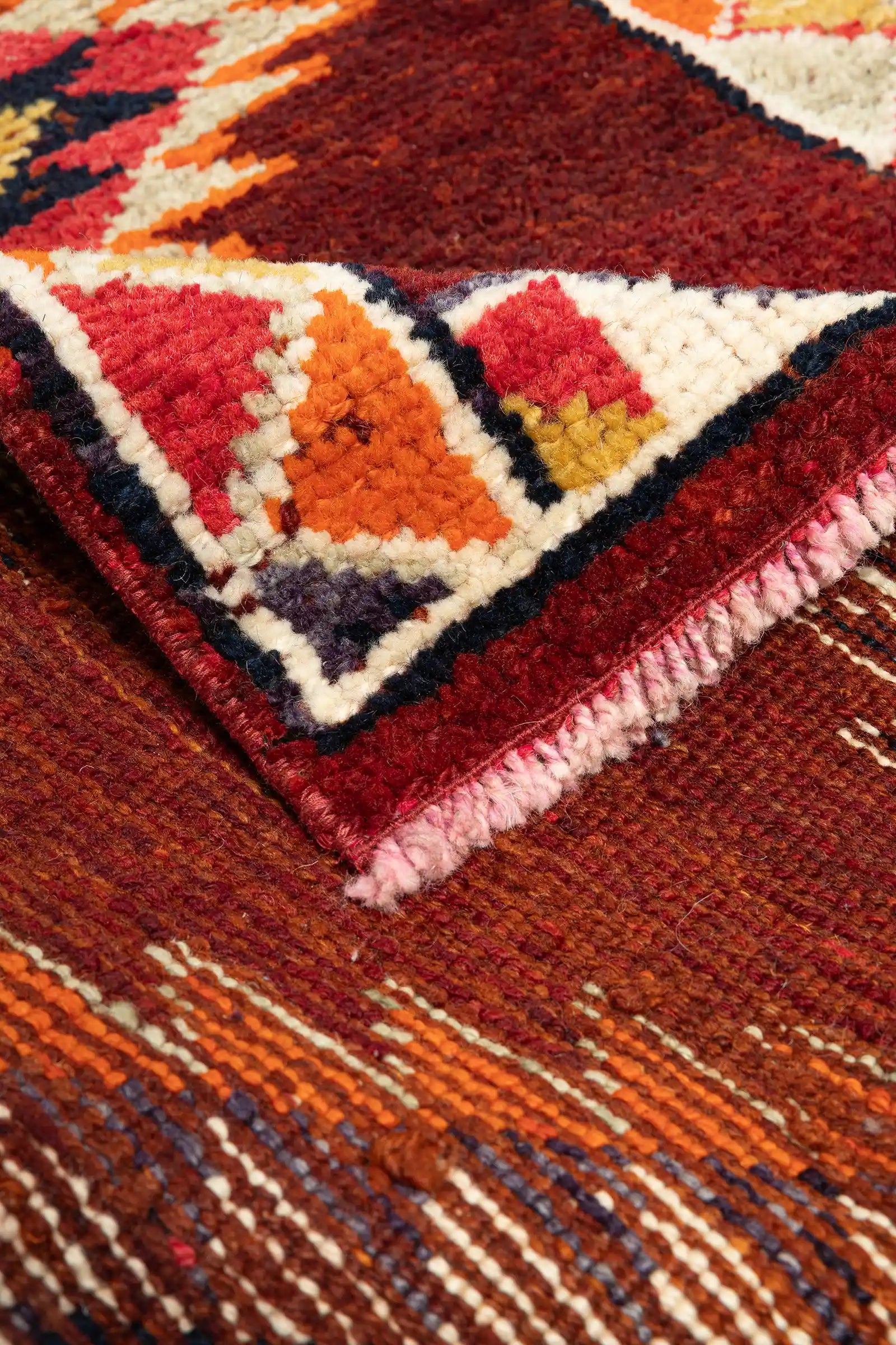 Detailansicht der umgeschlagenen Ecke des Teppichs, zeigt Fransenabschluss und Rückseite mit gestreifter Webung in Rot, Orange, Weiß und Braun.