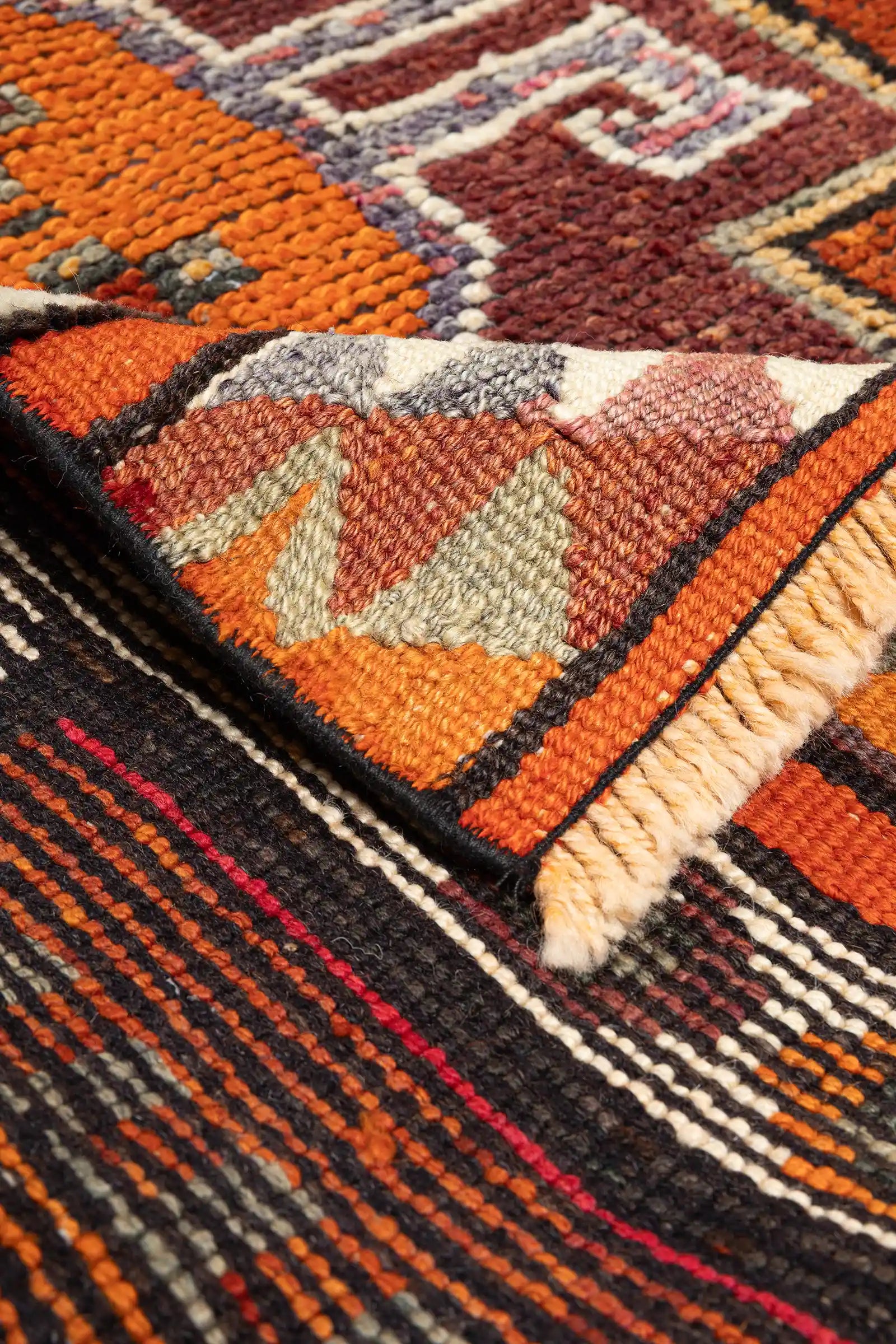 Detailaufnahme einer Teppichecke mit umgeschlagener Kante, sichtbar sind die bunte Rückseite in rot-schwarzer Streifenstruktur sowie helle Fransen am Abschluss.