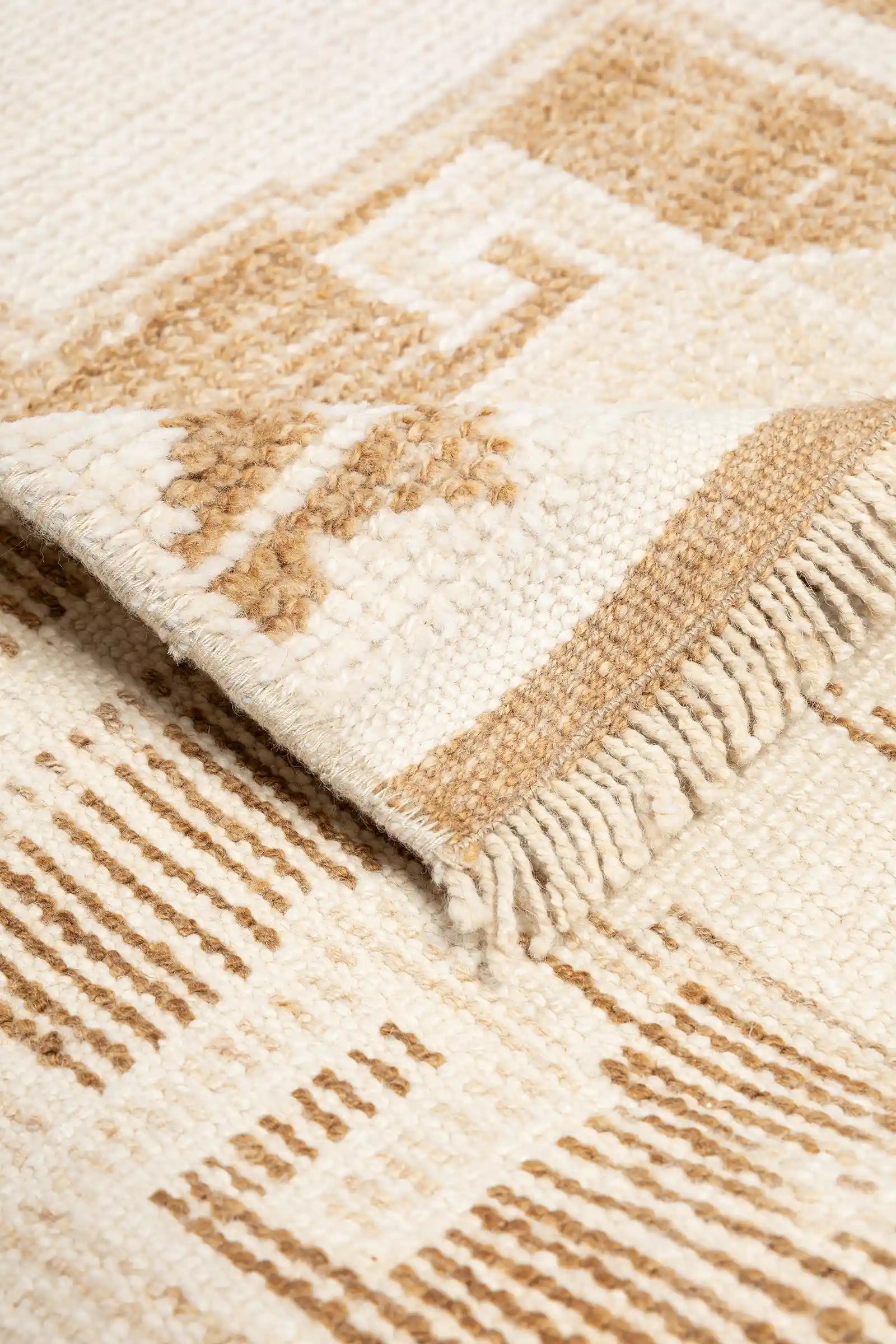 Detailaufnahme einer umgeschlagenen Ecke mit kurzen, geknoteten Fransen und gestreifter Rückseite in natürlichen Brauntönen auf cremefarbenem Grund.