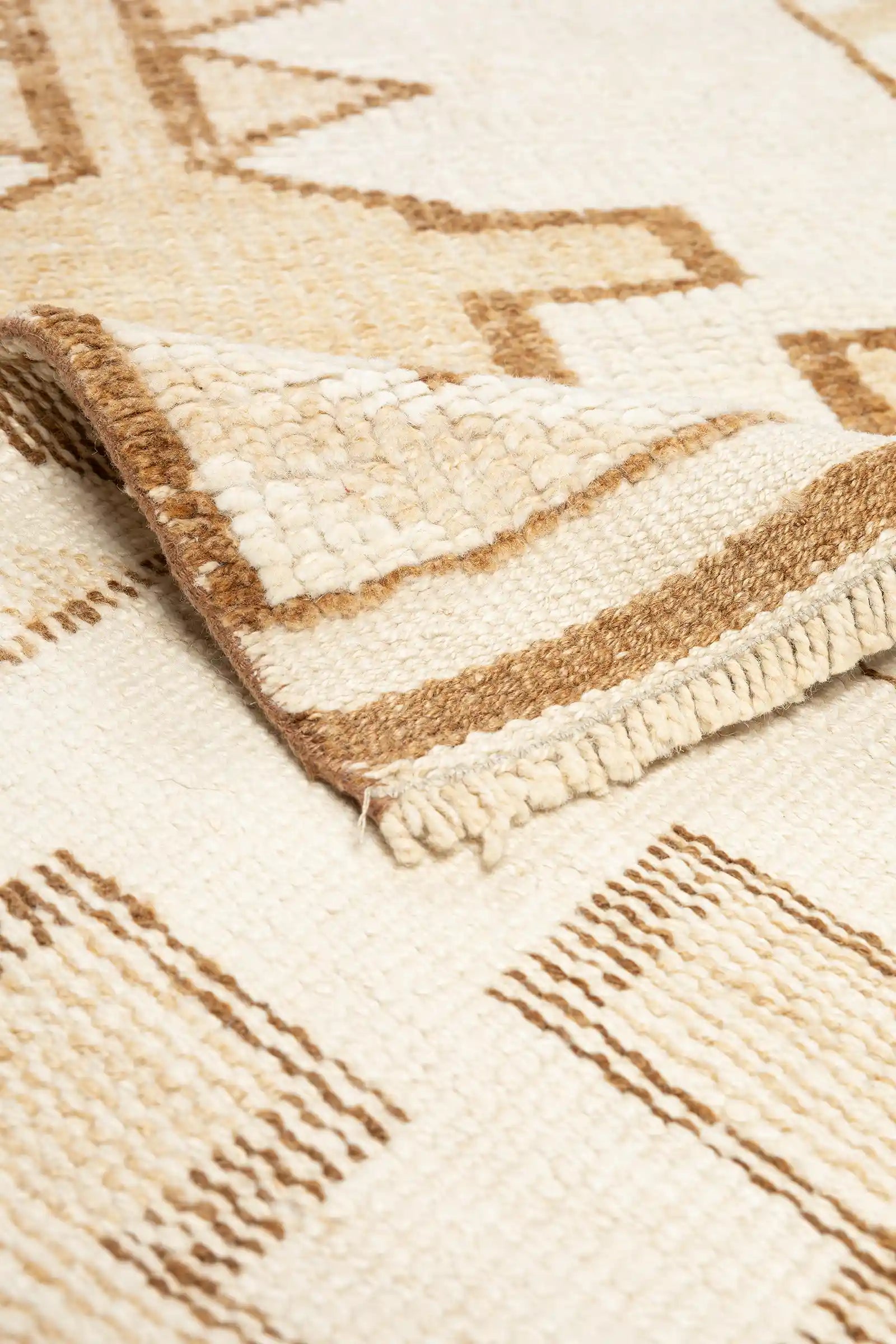 Detailansicht einer Teppichecke mit umgeschlagener Kante und sichtbarer Rückseite, zeigt gestreifte Musterung in Beige-Braun sowie kurze, dichte Fransen entlang der Kante.
