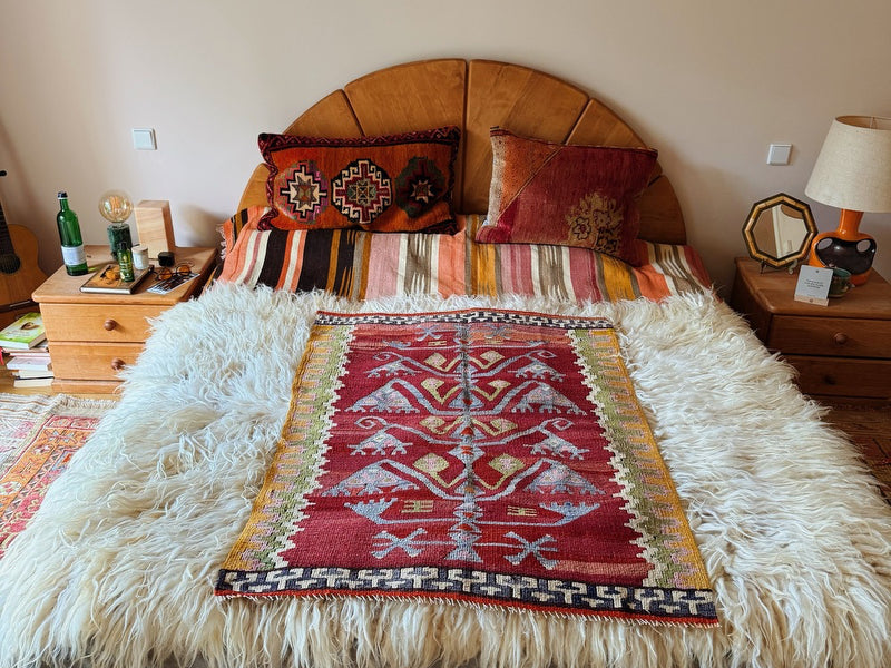 Holzbett mit vintage Kilim-Kissen, gestreiftem Läufer, langflorigem Schaffell und zentral platziertem anatolischen Teppich auf rotem Grund.