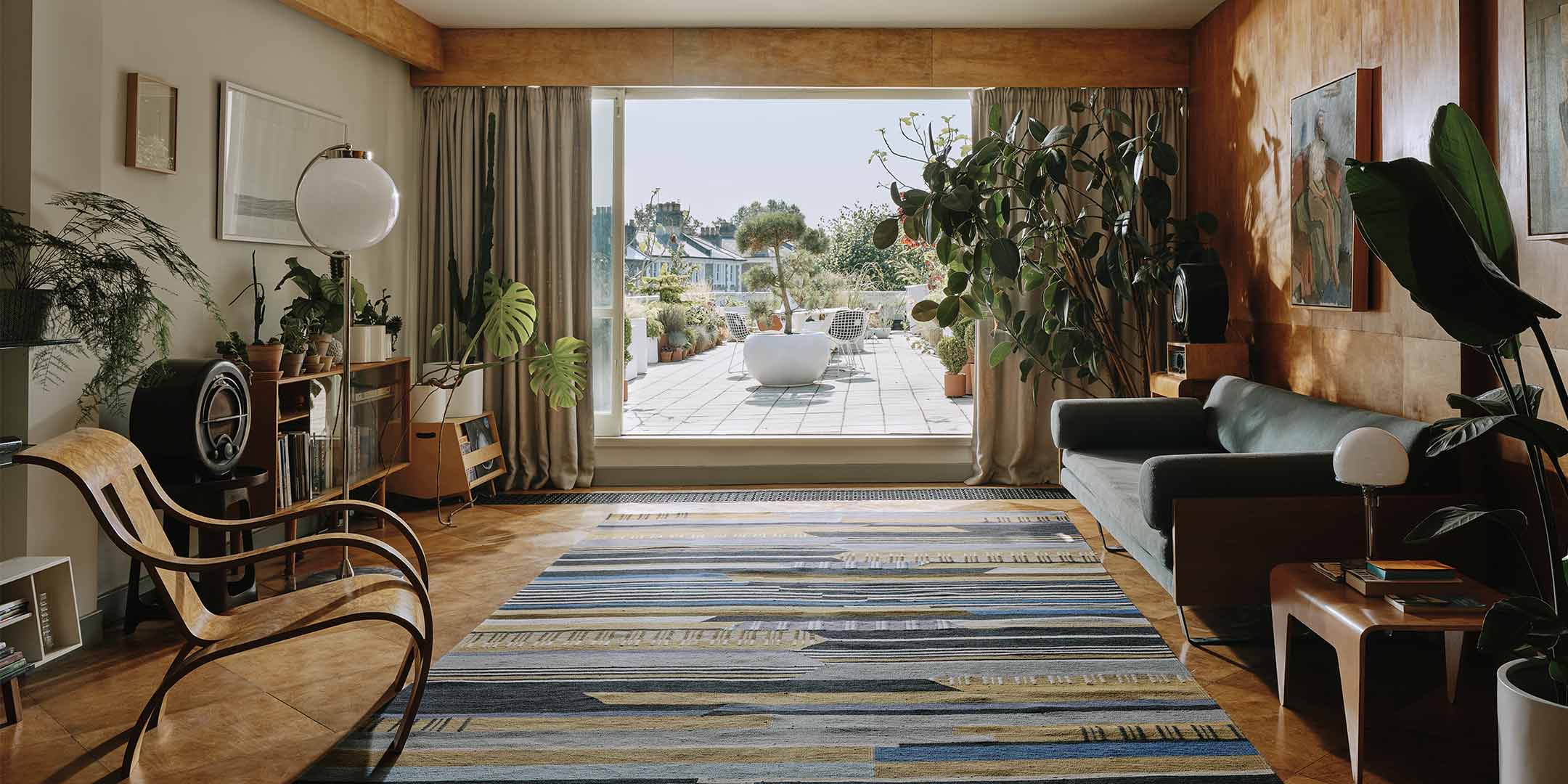 Großer Raum, der mit Pflanzen, Holzmöbeln und einem dunkel gestreiften Bauhaus Teppich eingerichtet ist und auf eine Terrasse hinaus führt