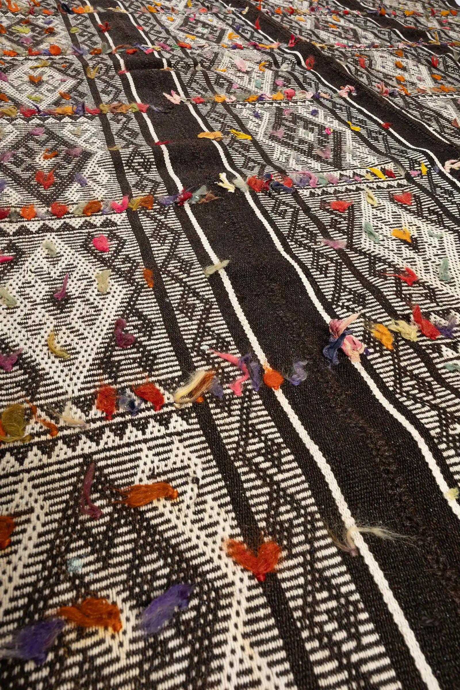 Extreme Nahaufnahme der Webstruktur mit kontrastierendem Schwarz-Weiß-Muster, durchsetzt von kleinen Garnbündeln in kräftigen Farben wie Orange, Lila und Rosa.