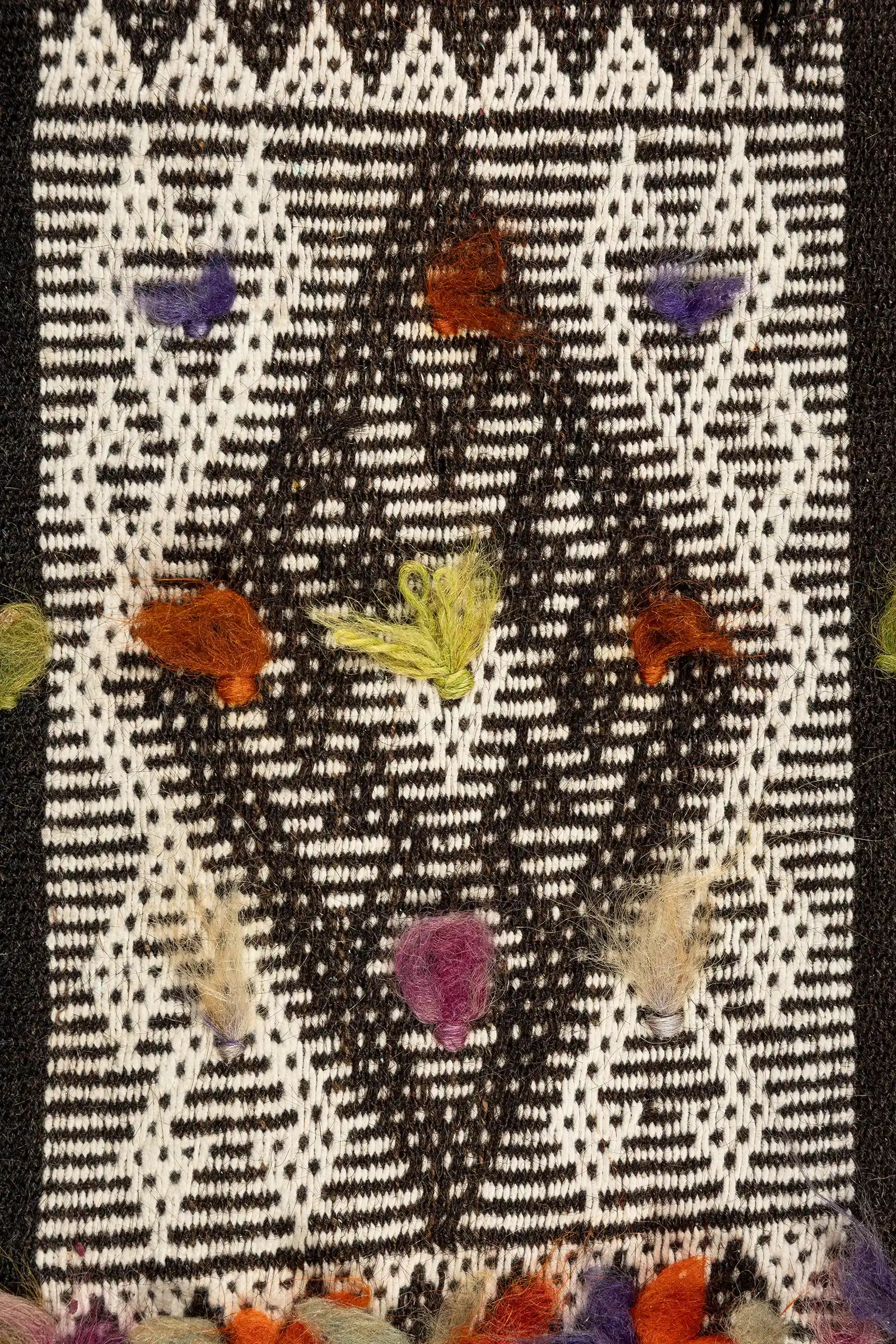 Nahansicht eines weißen Rautenmusters auf schwarzem Grund mit punktartigem Webmuster und vereinzelten bunten Garnknoten, darunter violett, orange, hellgrün und beige.