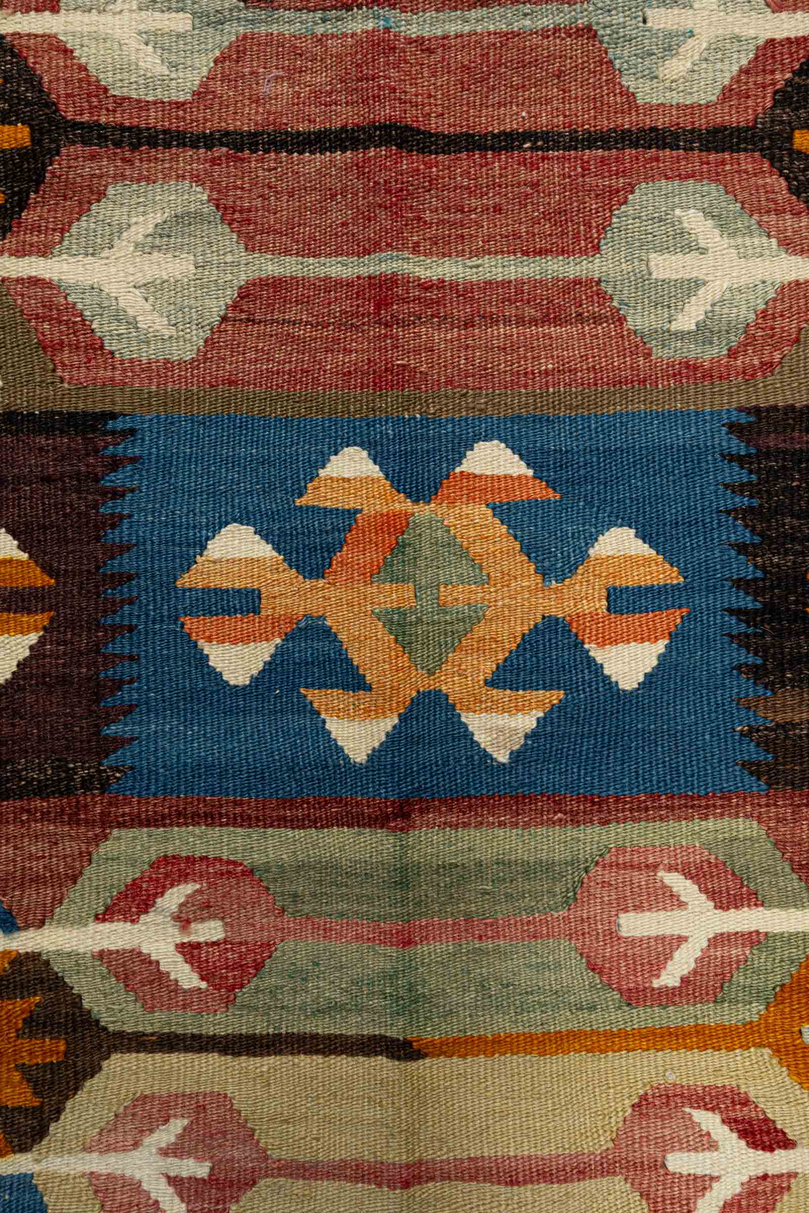 Nahe Aufnahme eines Streifenabschnitts mit blauen, schwarzen und senfgelben geometrischen Mustern auf rotbraunem Grund, ergänzt durch cremefarbene, pfeilartige Ornamente und einen olivgrünen Rand mit rosa Elementen.

