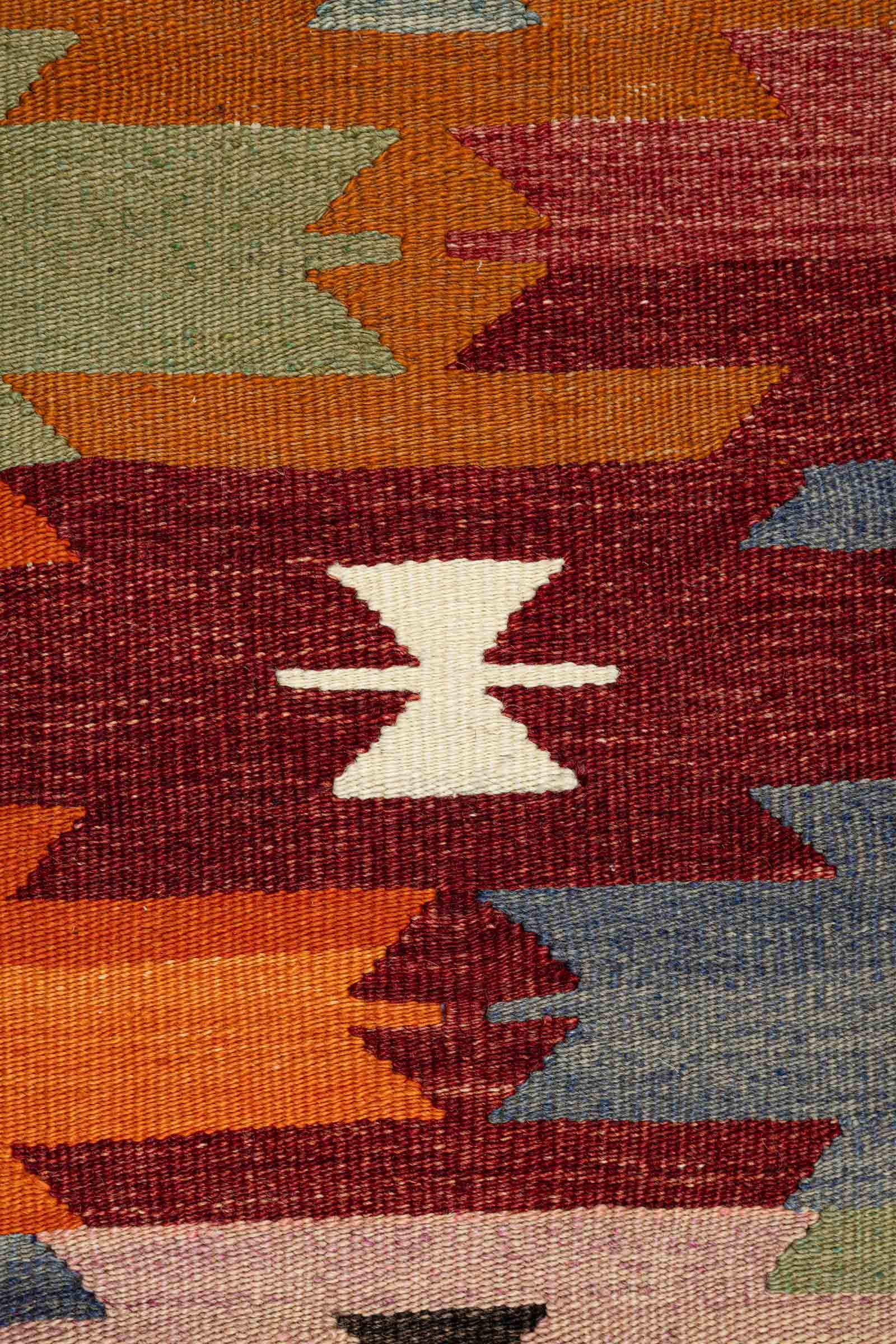 Nahansicht eines cremeweißen geometrischen Symbols in Sanduhrform auf dunkelrotem Grund, umgeben von spitz zulaufenden Zacken in orange, olivgrün, weinrot und blau auf dichter, flachgewebter Oberfläche.
