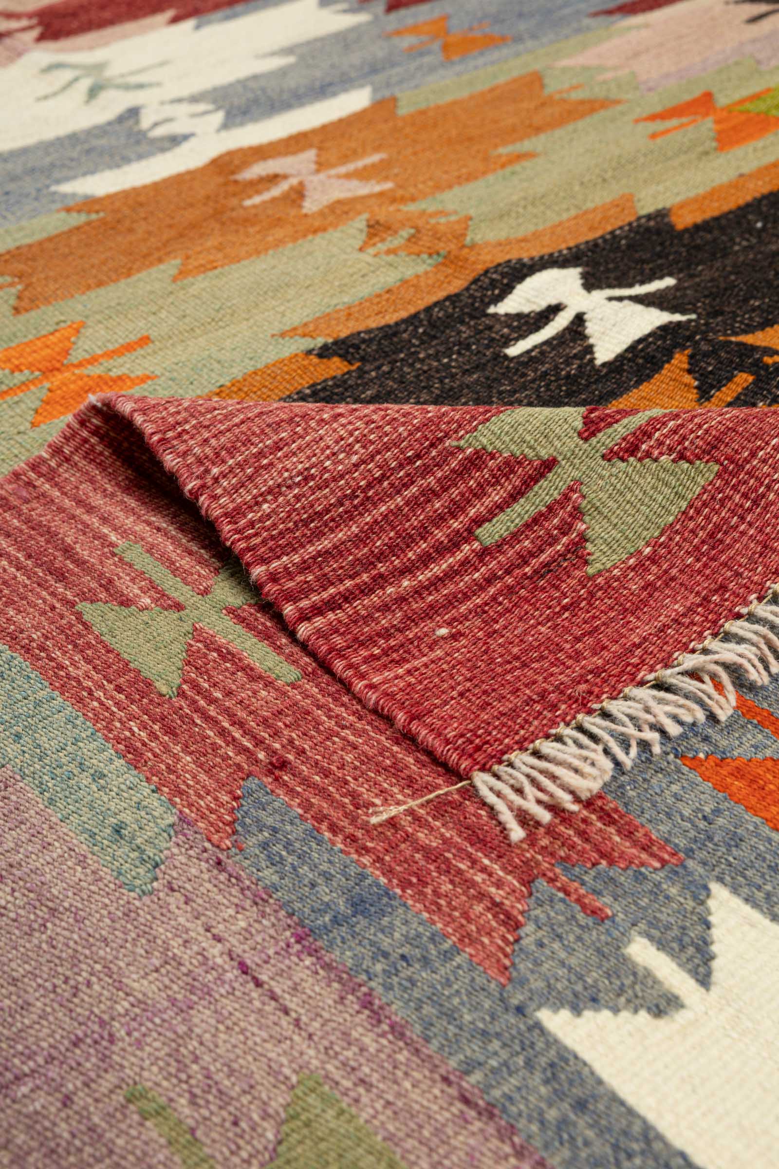 Ansicht einer Ecke des Kilims mit umgeschlagener Kante, zeigt die rot melierte Rückseite mit grünem Muster und weißen Fransen am Rand sowie die fein gewebte Vorderseite mit schwarzen und orangen Symbolen.