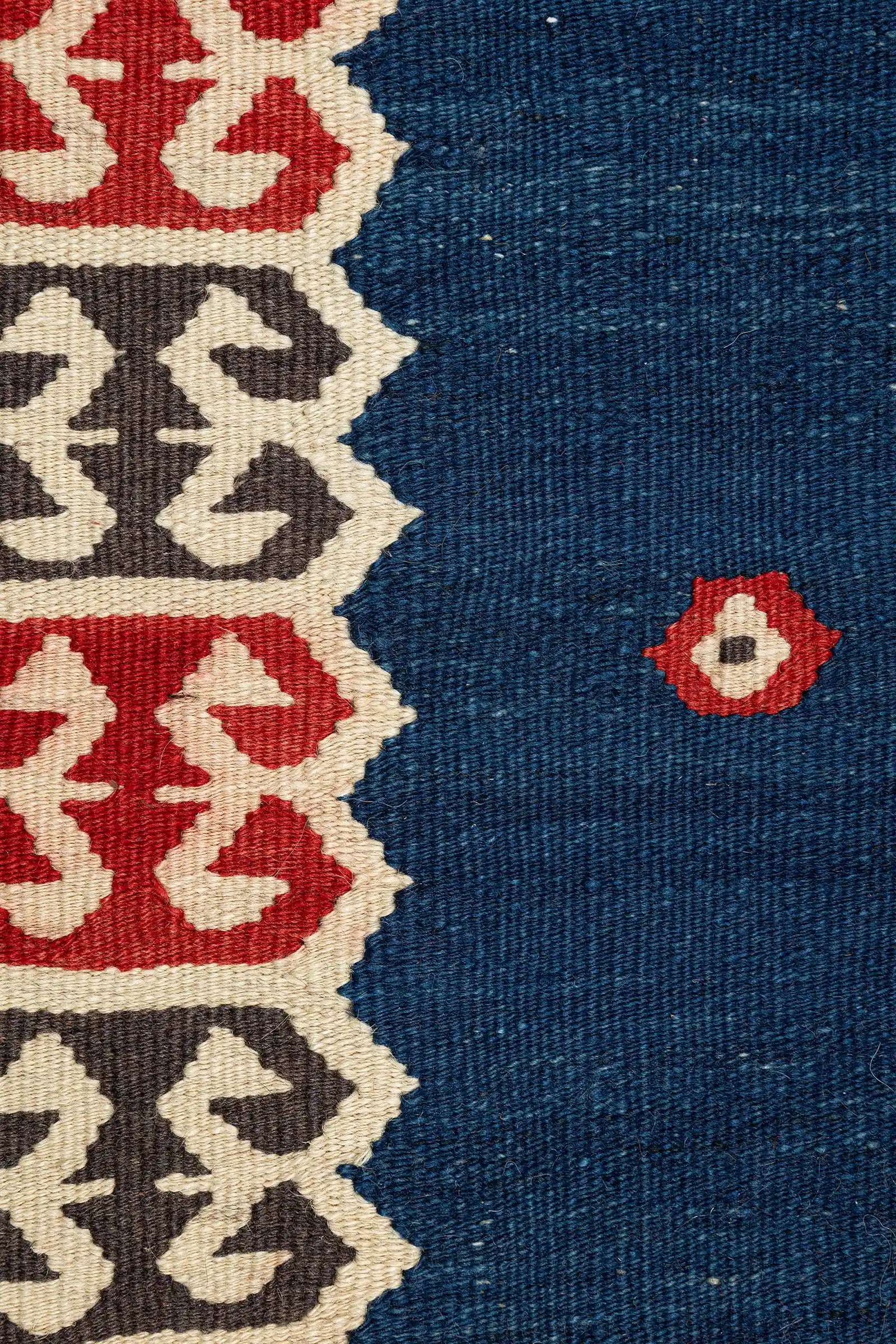 Nahaufnahme des blauen Kilimbereichs mit eingewebten roten, blütenförmigen Akzenten und kontrastierender Bordüre mit roten und braunen Ornamenten auf beigem Grund.
