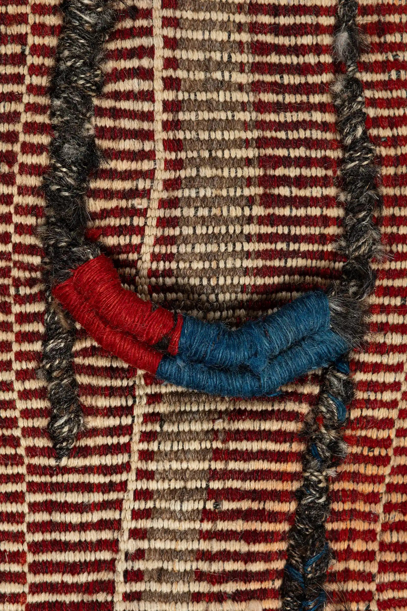 Nahansicht der fein gewebten Textur in rostroten und cremefarbenen Querstreifen mit dunkler Vertikalnaht. Deutlich erkennbar ist eine schwarze Schnur mit fester blauer und roter Umwicklung sowie grobe Garnverdickungen.
