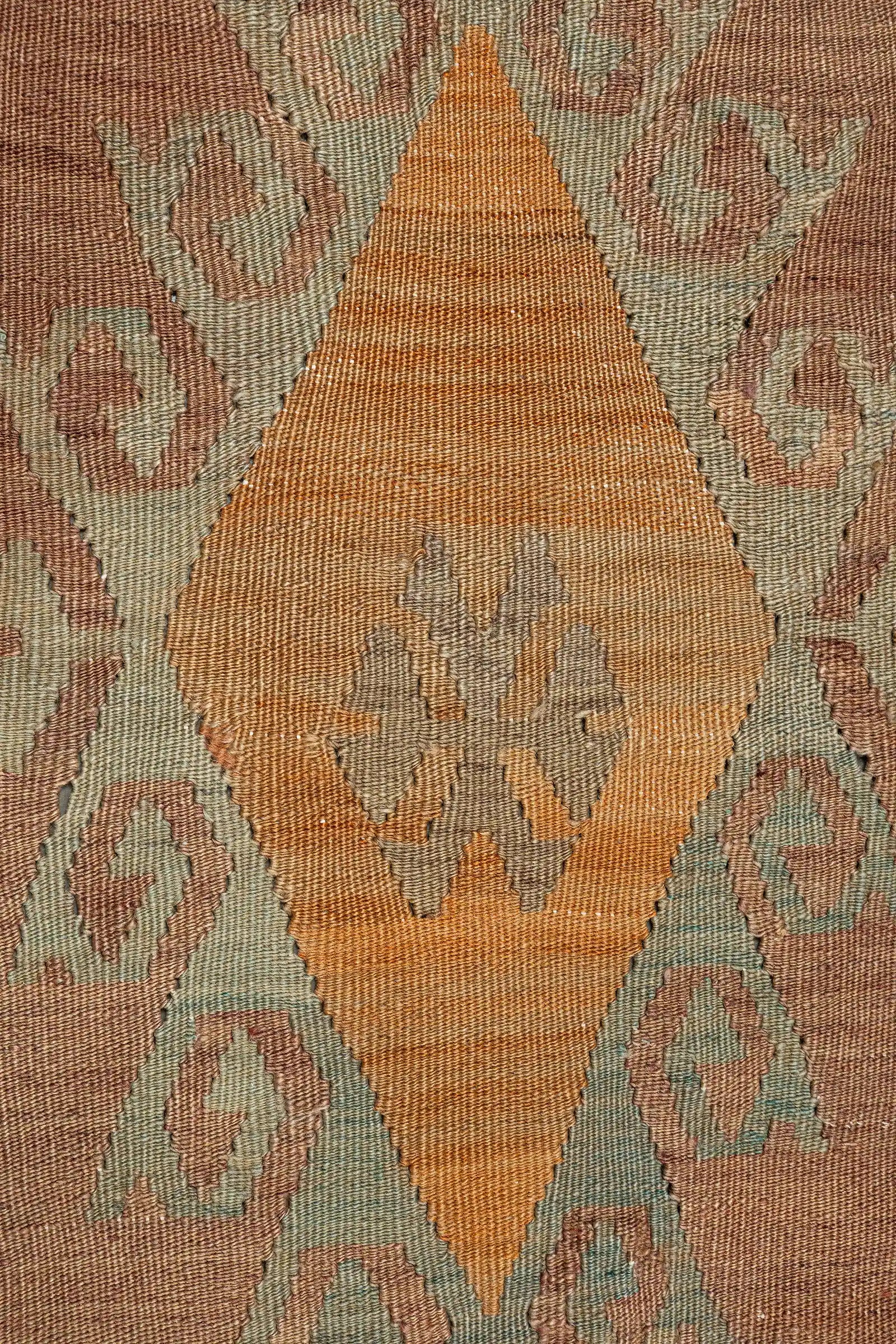 Nahaufnahme eines ockerfarbenen, rautenförmigen Zentralmotivs mit symmetrischem Muster in Blaugrau und Beige, eingebettet in ein Umfeld aus hellen, ornamentalen Elementen auf rosafarbenem Grund.